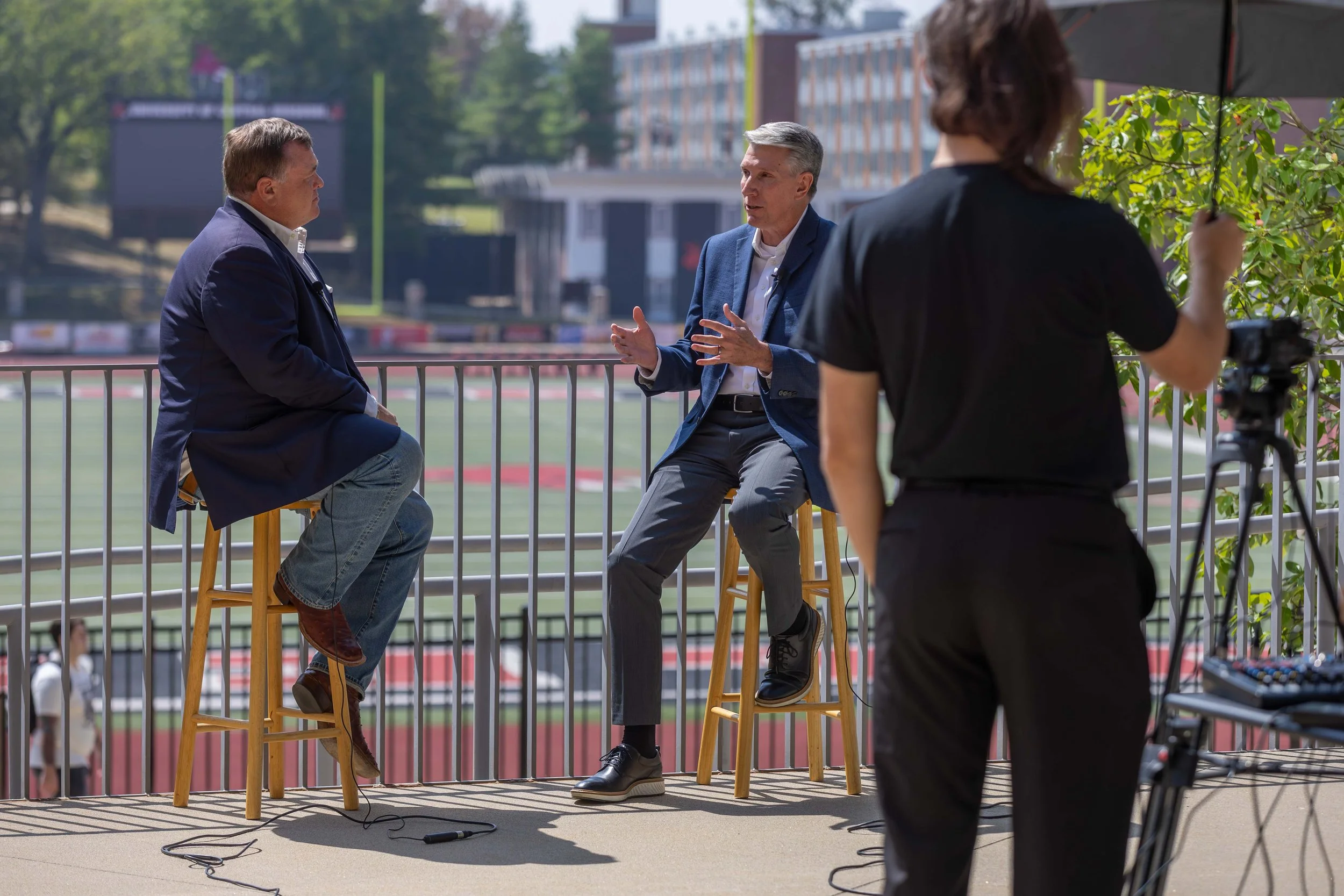 Two men in business attire are seated on stools and engaged in conversation on an outdoor balcony or deck, with a woman operating camera and equipment in the foreground, and a sports field with a running track and a building in the background.