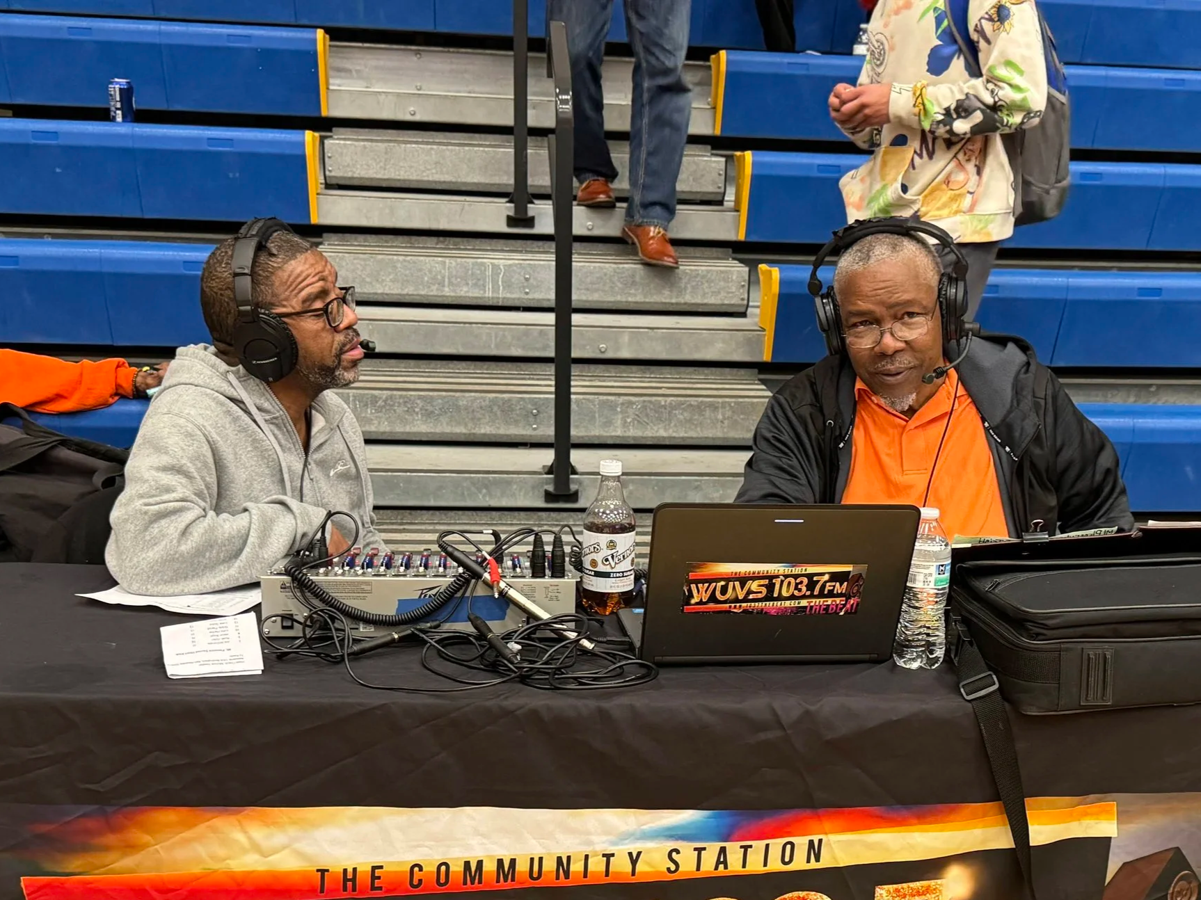 Two men wearing headphones and microphones sitting at a table with electronic equipment and a laptop, broadcasting at a community event in a gymnasium with blue bleachers in the background.