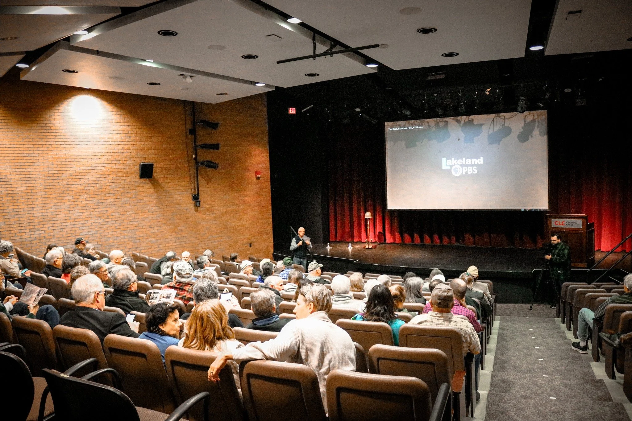 Audience seated in an auditorium, watching a presentation on stage with a large screen displaying the Lakeland PBS logo. A person is speaking at the front, and a camera operator records the event.