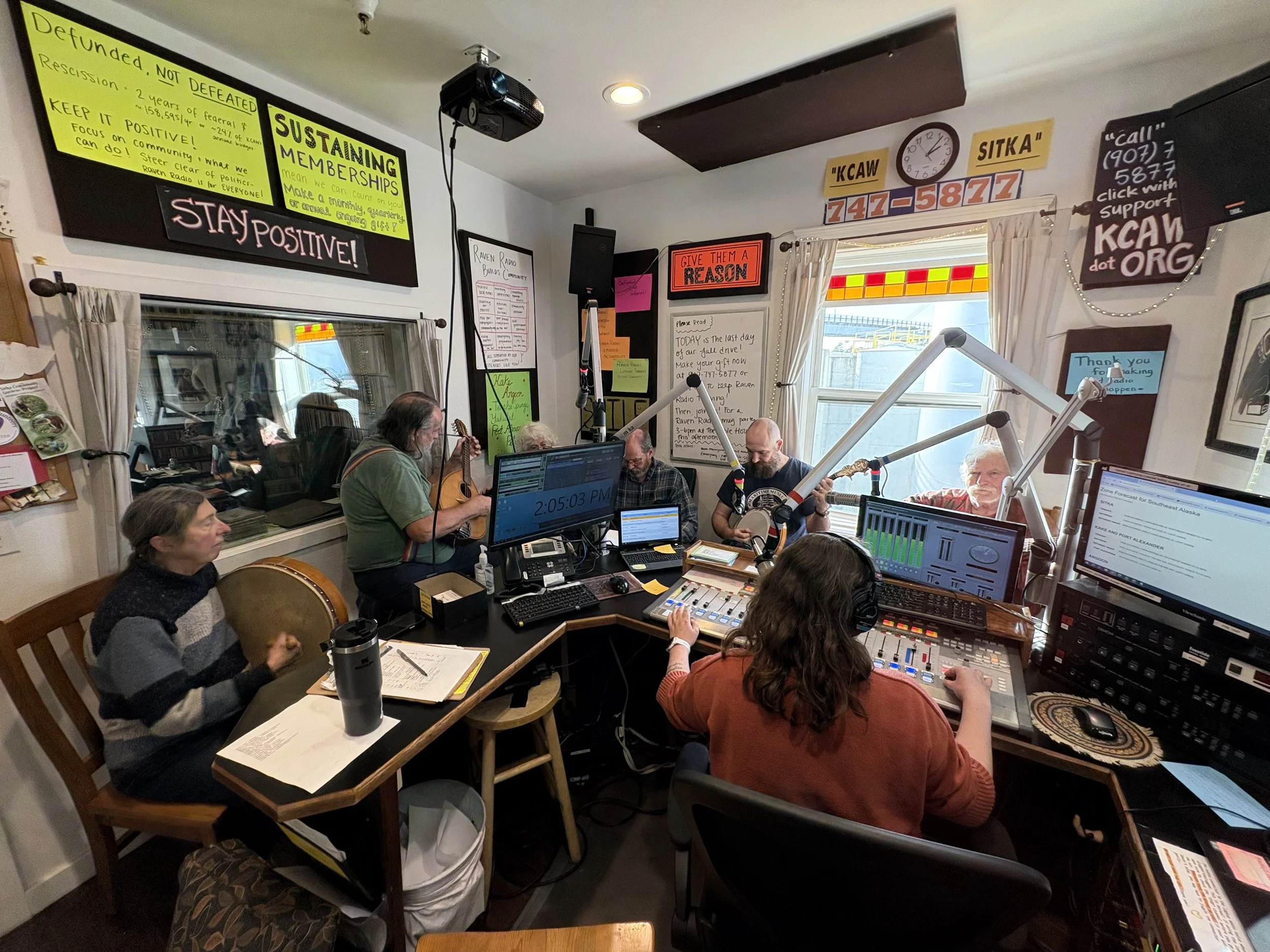 People inside a radio studio with musical instruments, broadcast equipment, and multiple monitors, some playing guitars, during a live recording or broadcast.
