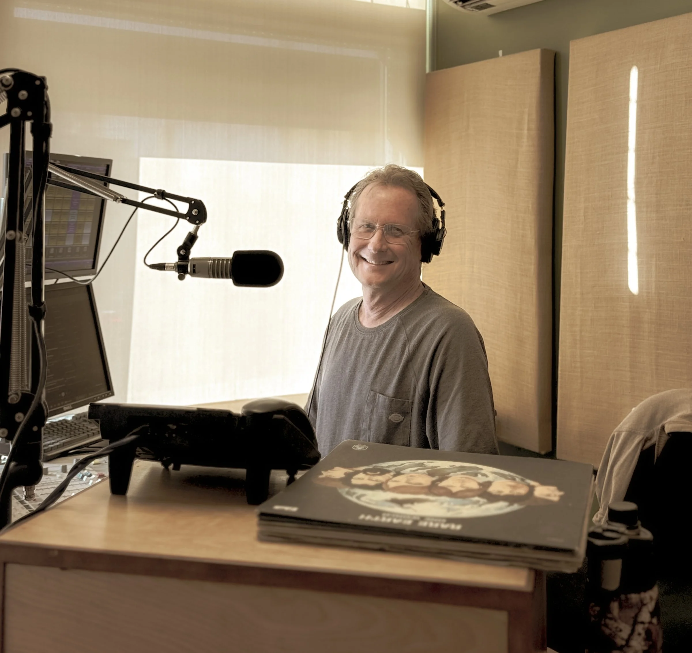 A man sitting at a radio station or recording studio with headphones, smiling at the camera. There are microphones, computer monitors, and a stack of magazines or records on the desk.