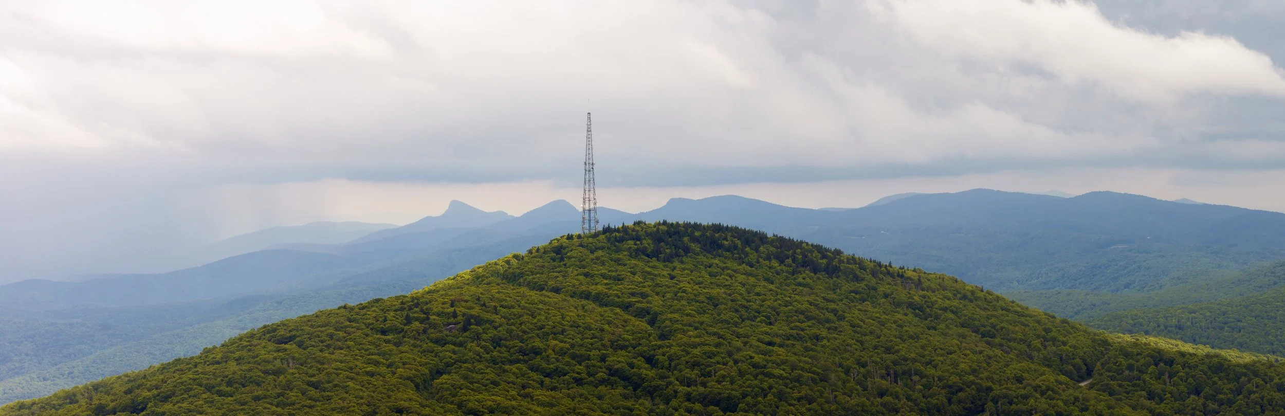 A green, forested mountain with a communication tower at its peak and mountain ranges in the background under a cloudy sky.