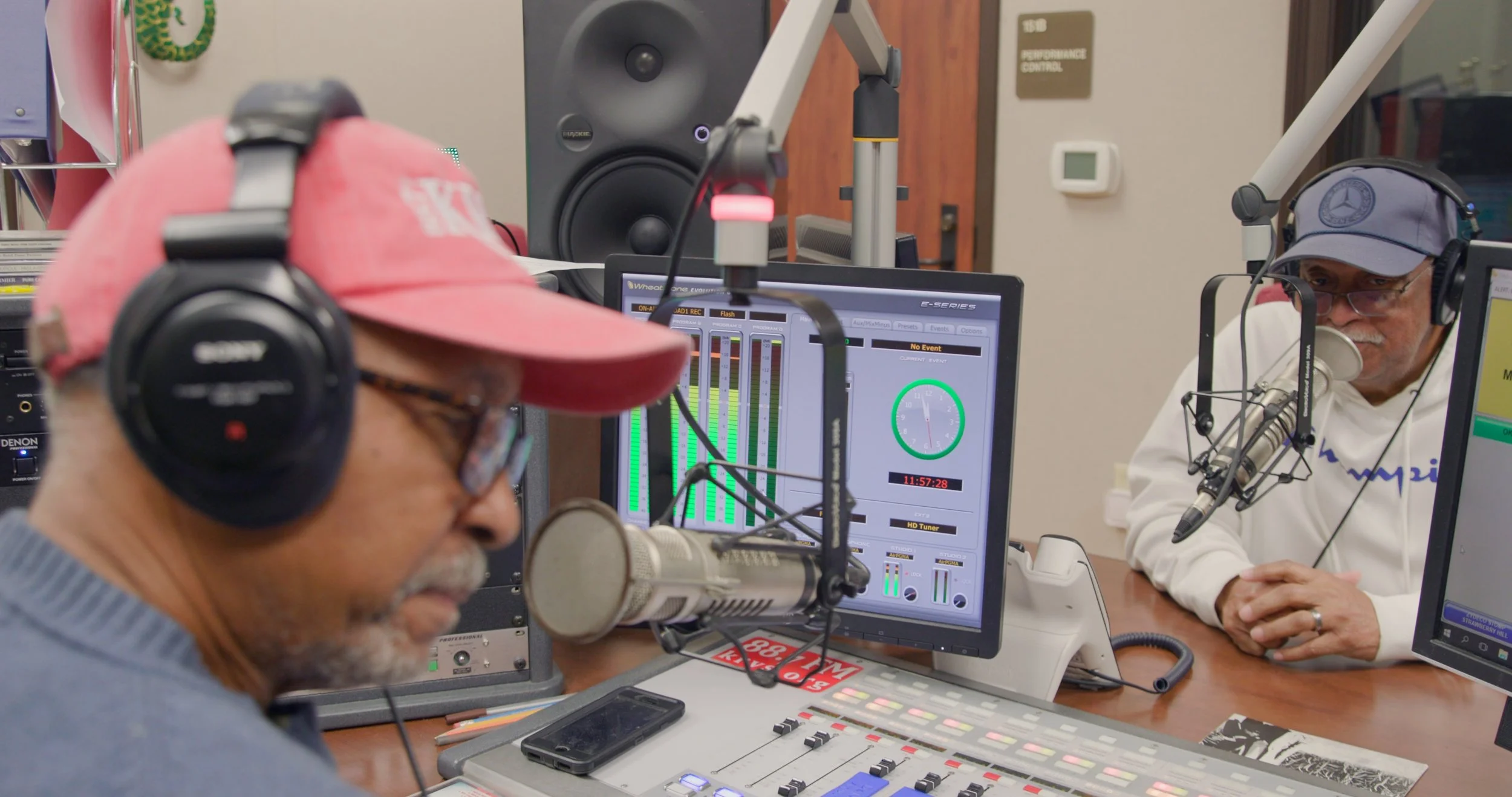 Two men in baseball caps and over-the-ear headphones are pictured sitting in a radio studio with microphones, a control board, and computer screens in front of them.