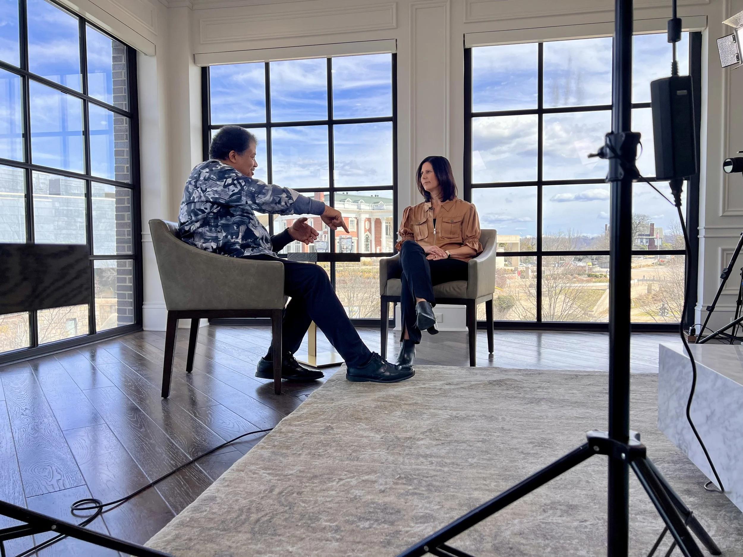 Two women having a conversation in an interview setting in a bright room with large windows and city view.