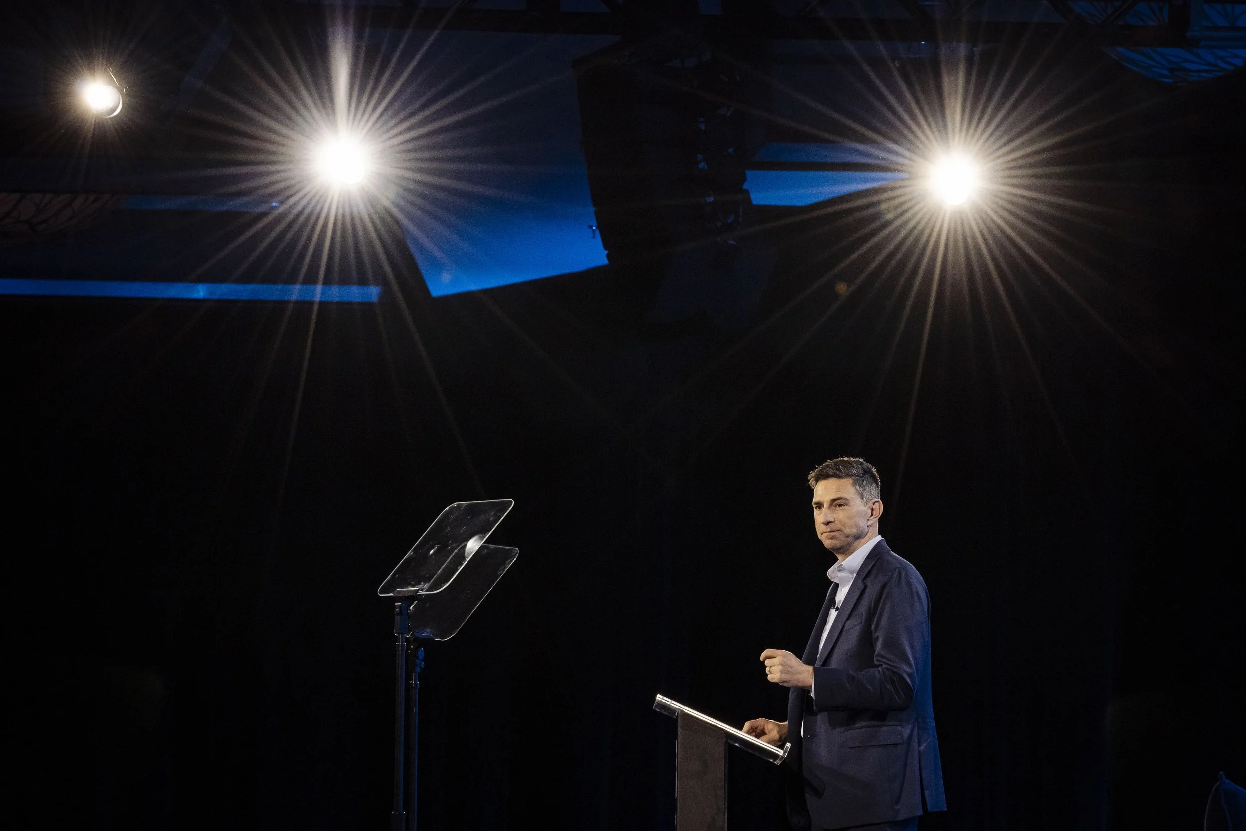 A man in a dark suit speaks from a podium against a dark blue backdrop with spotlights in the background.