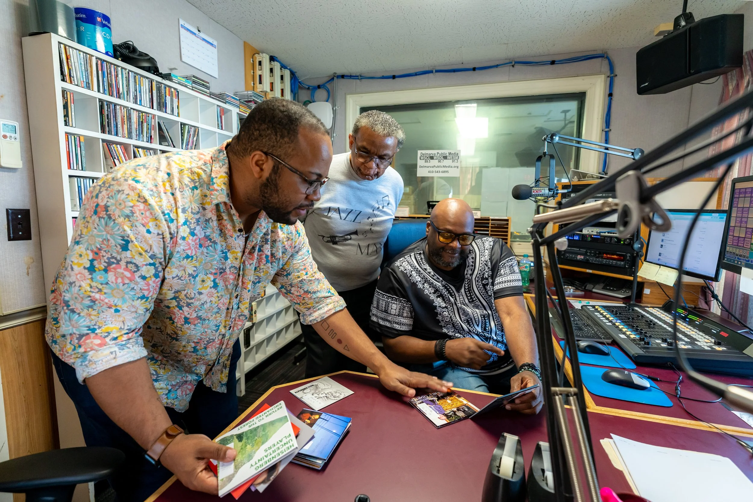 Three men huddle around broadcasting equipment in a radio studio, with a broadcast microphone and audio mixing board in the foreground.