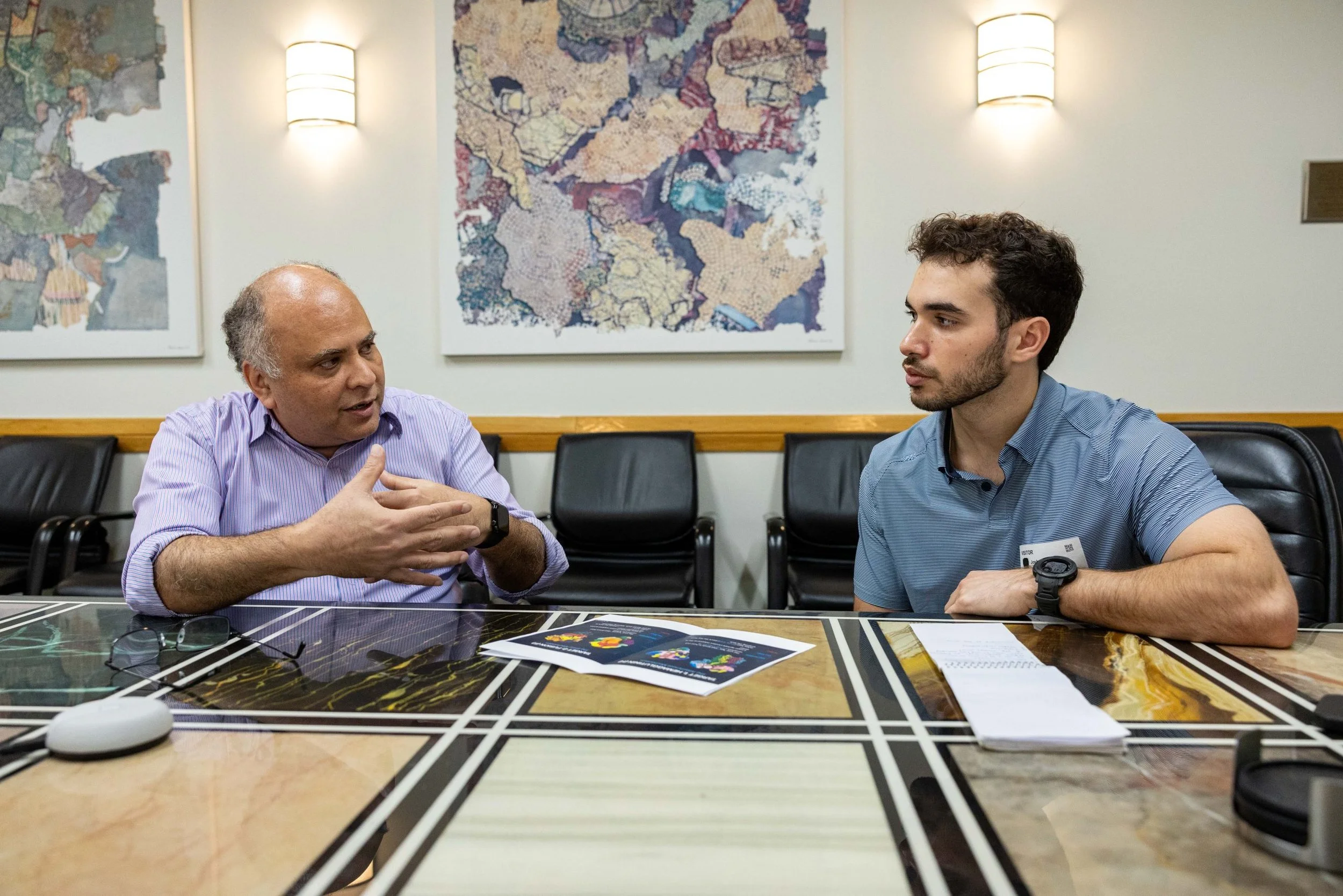 Two men engaged in conversation at a multicolored conference table, with a colorful abstract artwork on the wall behind them.