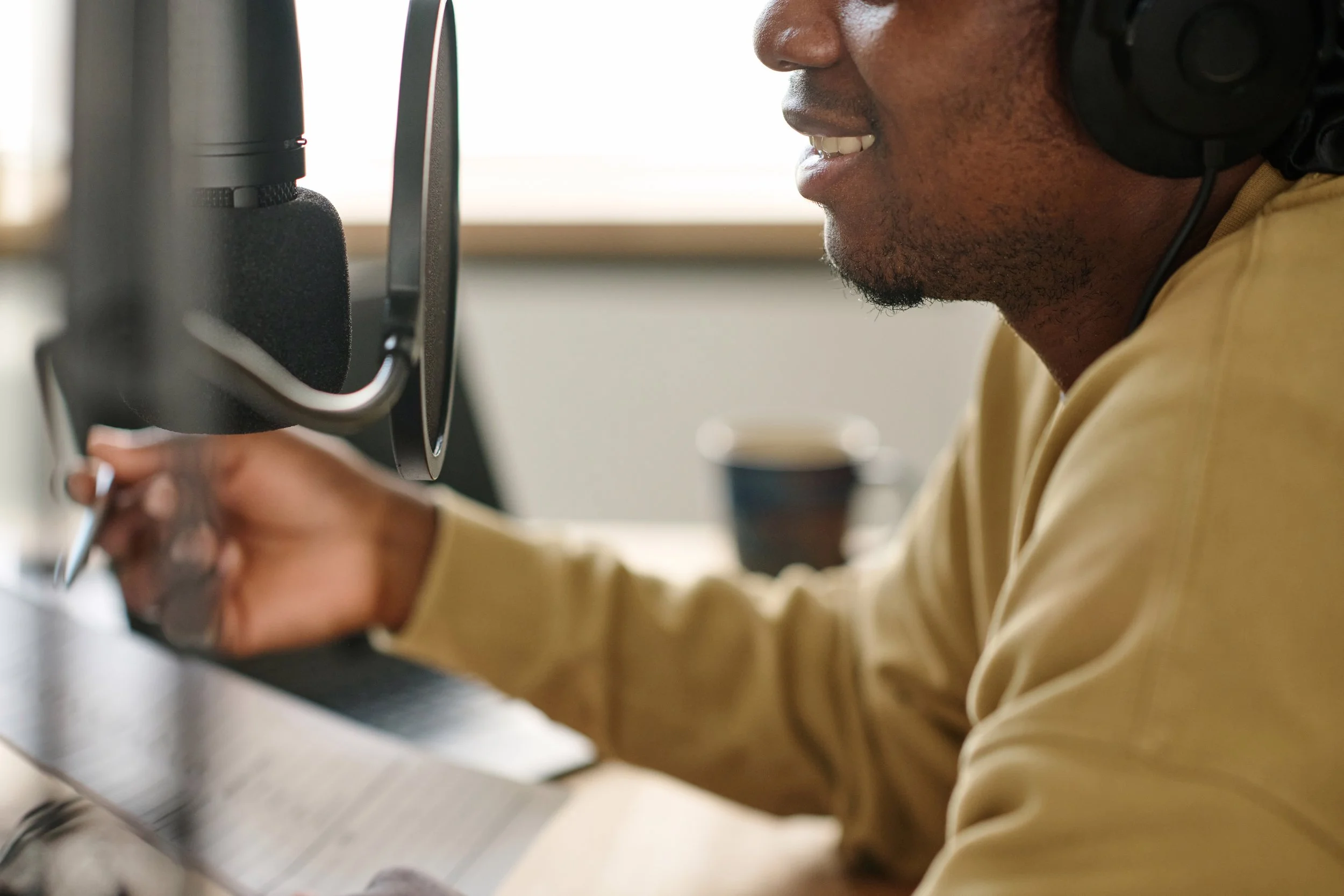 A person in a beige shirt recording in a professional home studio, wearing large over-ear headphones, sitting at a desk with a microphone, and writing in a notebook.