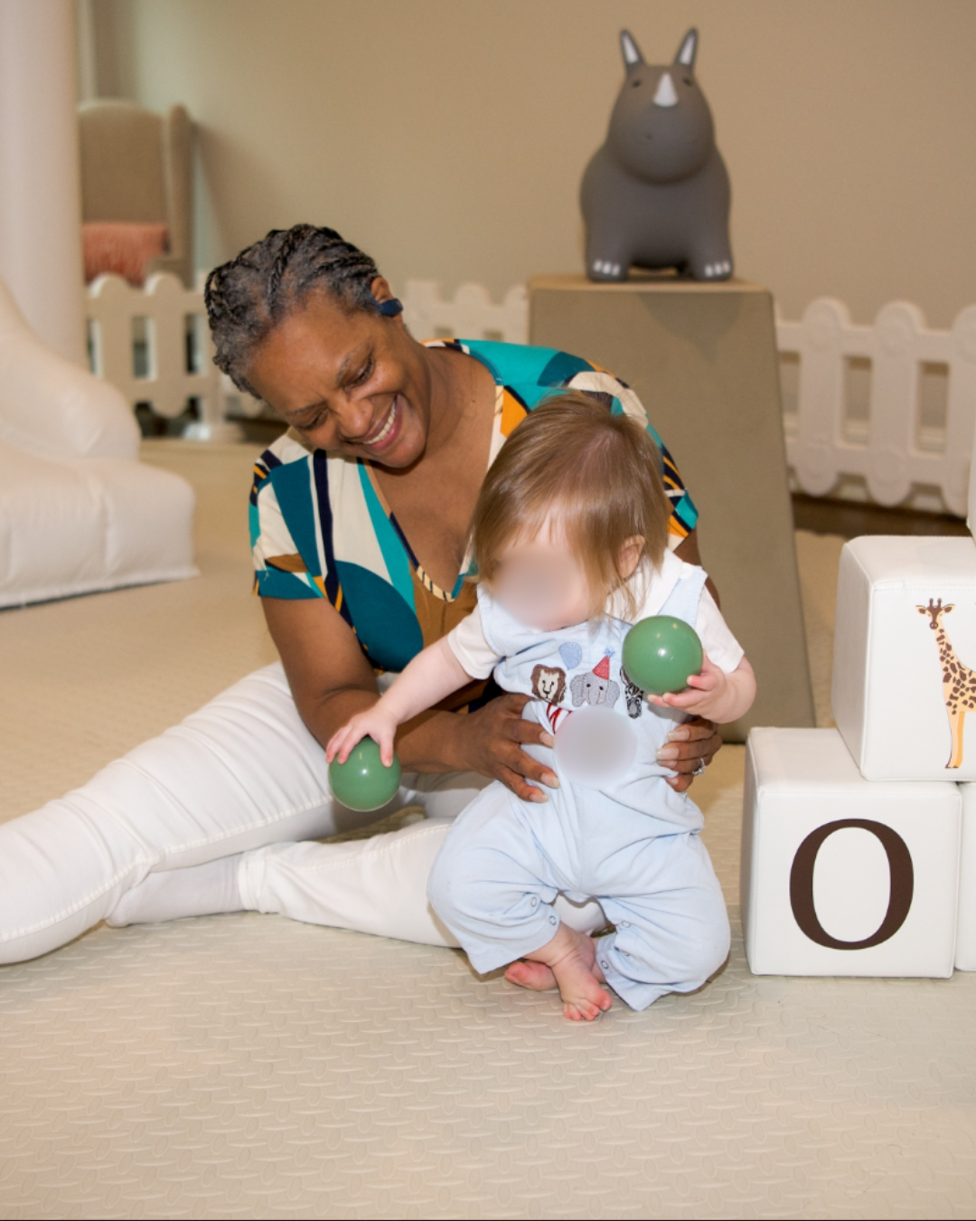 Erica Harris sitting on the floor smiling and playing with a baby holding soft toys in a nursery play space