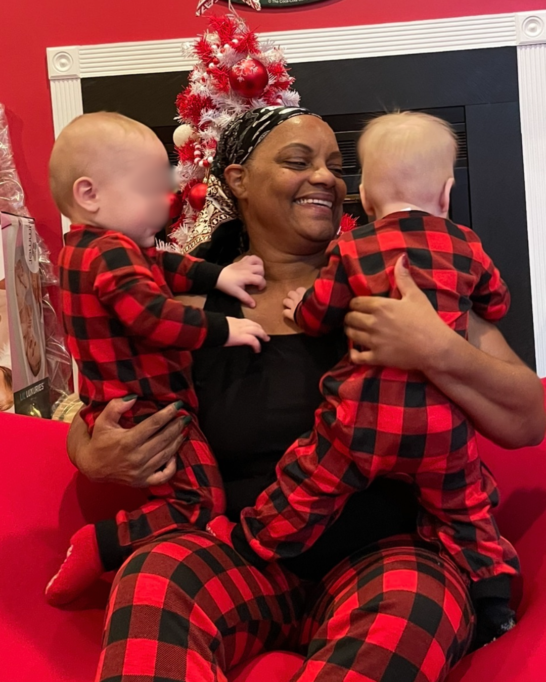 Erica Harris smiles while holding twin toddlers in matching red-and-black plaid pajamas in front of a Christmas tree