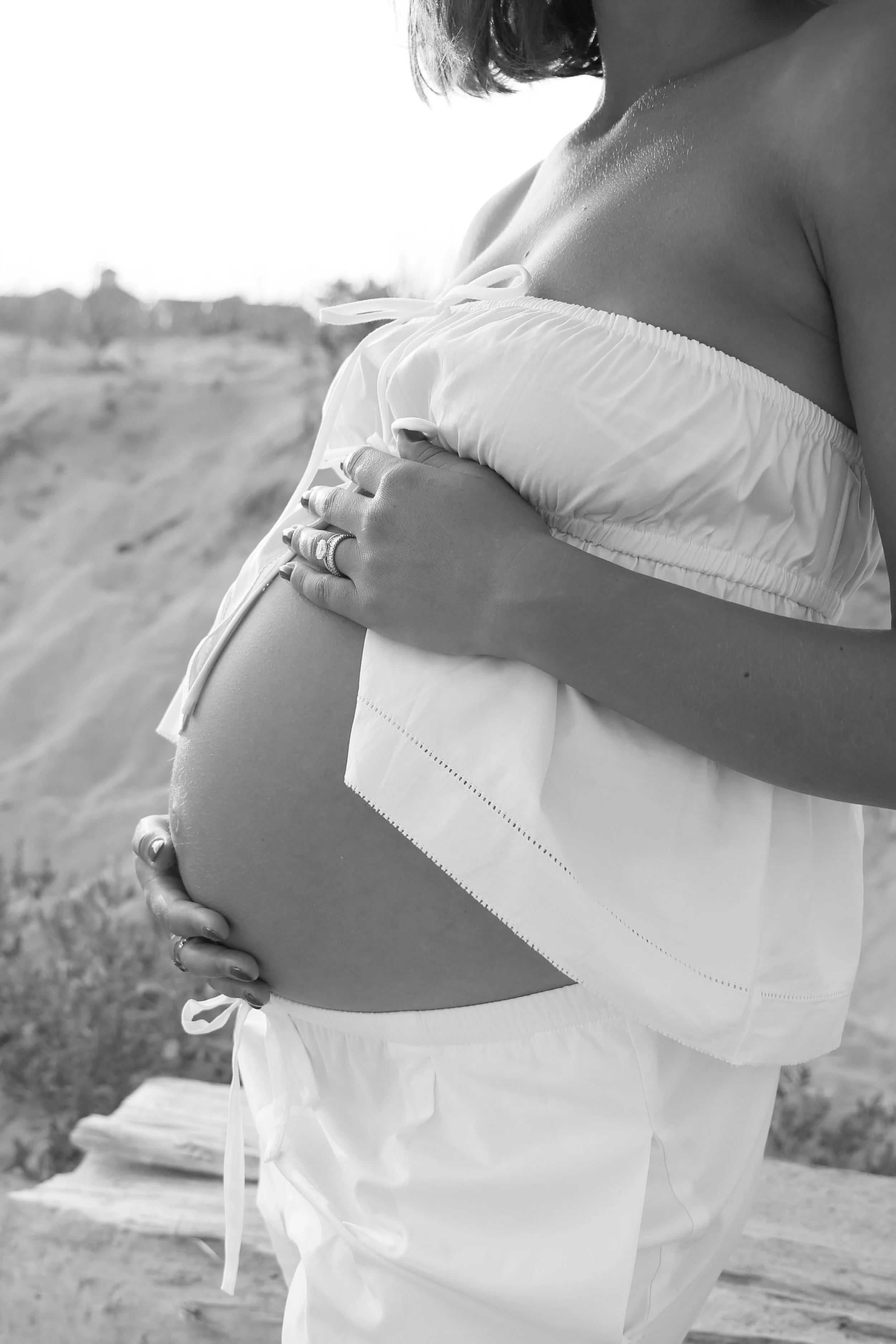 A pregnant woman with dark hair in a sleeveless top, gently holding her baby bump outdoors, in black and white.