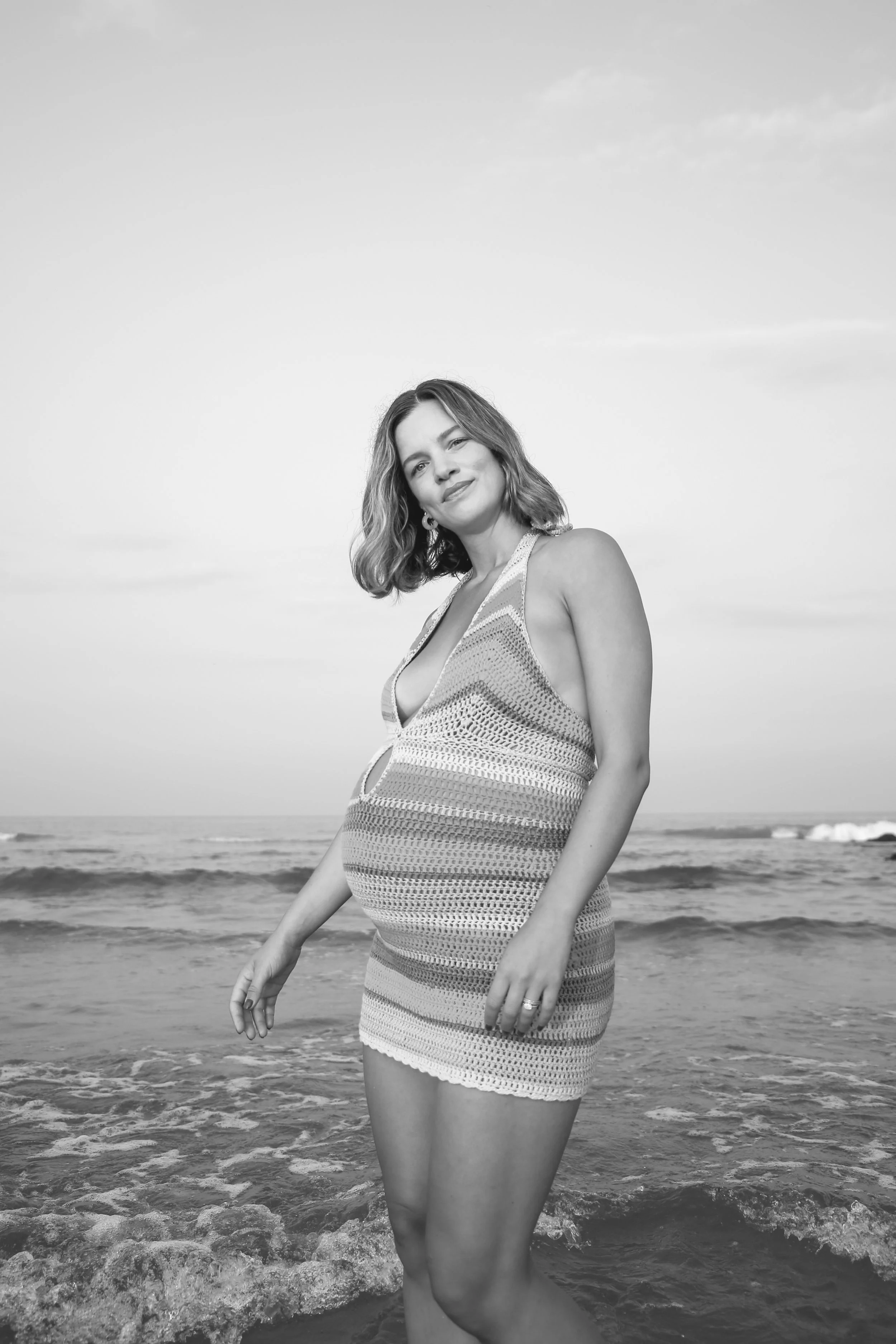 Black and white photo of a pregnant woman standing in the ocean with waves, wearing a sleeveless crochet dress and winking at the camera.