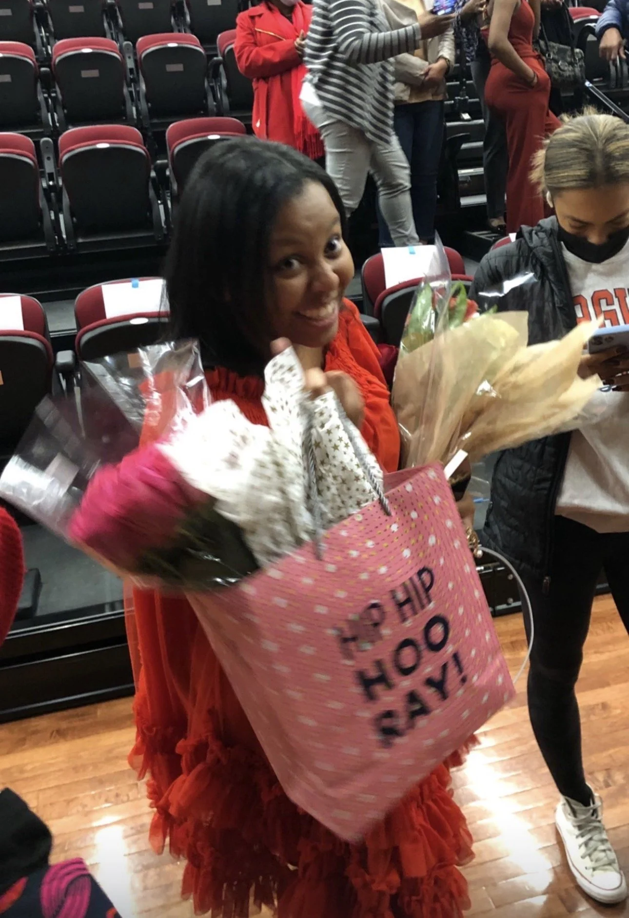 A woman smiling and holding bouquets of flowers and a pink gift bag with 'Happy Happy Happy' written on it, at an indoor event with empty seats in the background.