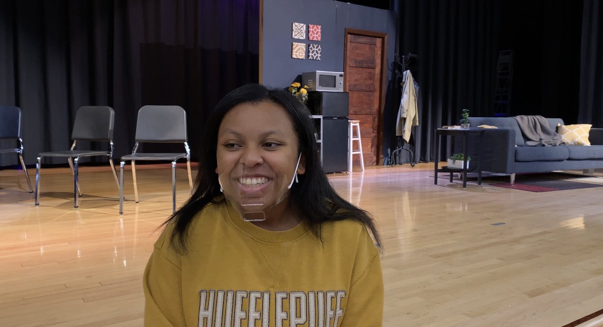 A woman with a smile wearing a yellow shirt with the word "HUFFLEPUFF" on it, sitting in a room with a stage background, chairs, a couch, and various decorations.