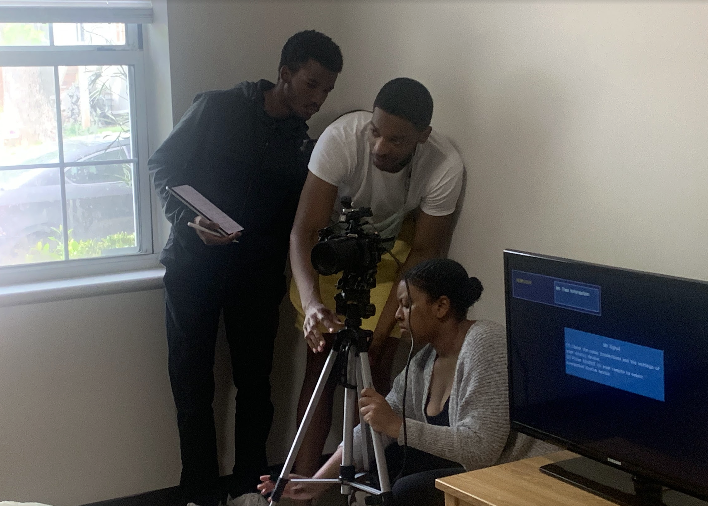 Four people, two men and two women, working with a camera and tripod in a room near a window. One woman is sitting, wearing headphones, and looking at the camera, while one man adjusts the camera and the other man holds a tablet.