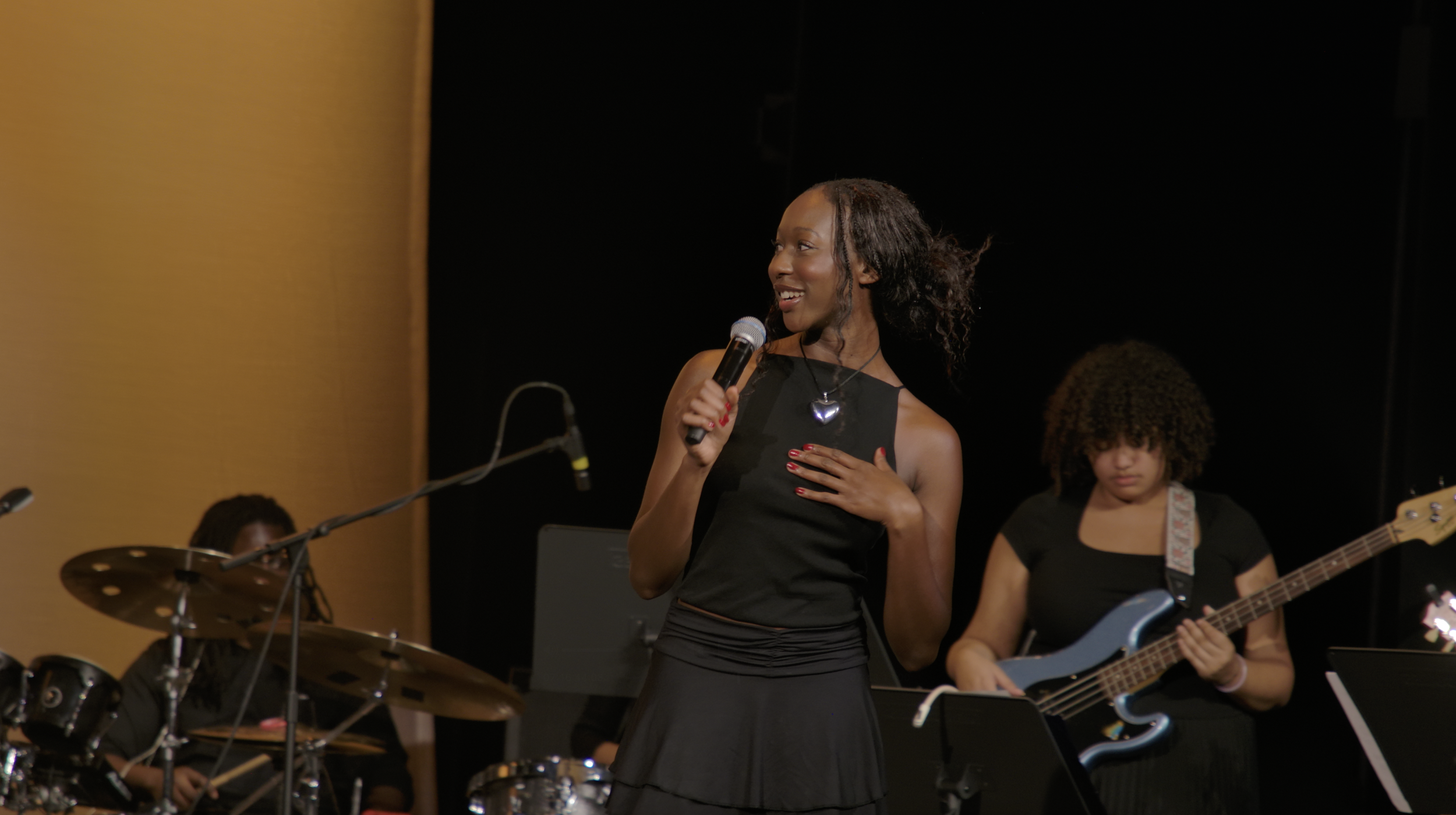 A woman singing into a microphone on stage, with a bass guitarist and drummer in the background.