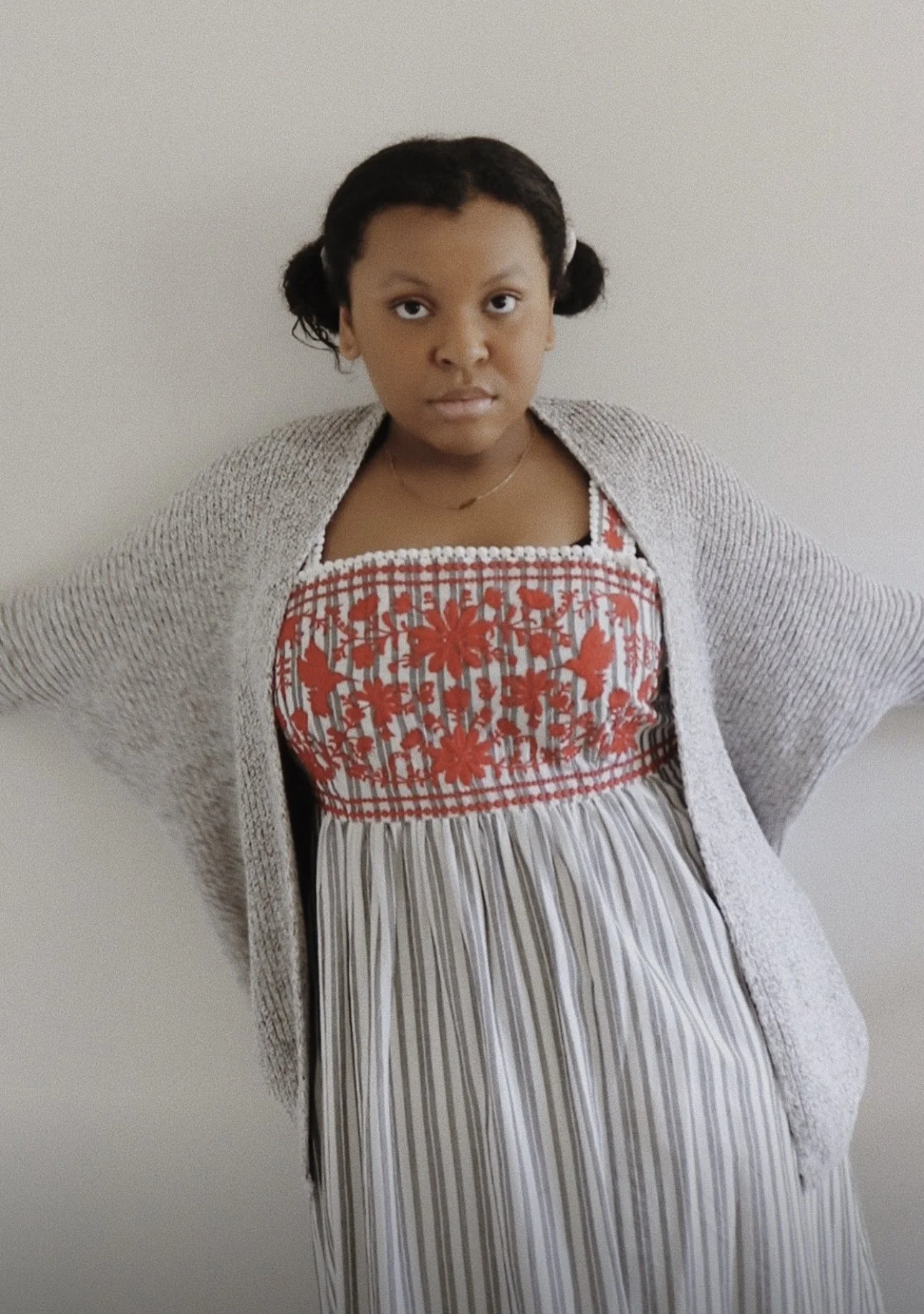 A young girl with dark hair styled in two buns, wearing a beige and white knitted cardigan over a colorful patterned dress with red floral and butterfly embroidery, standing against a plain, light-colored wall.