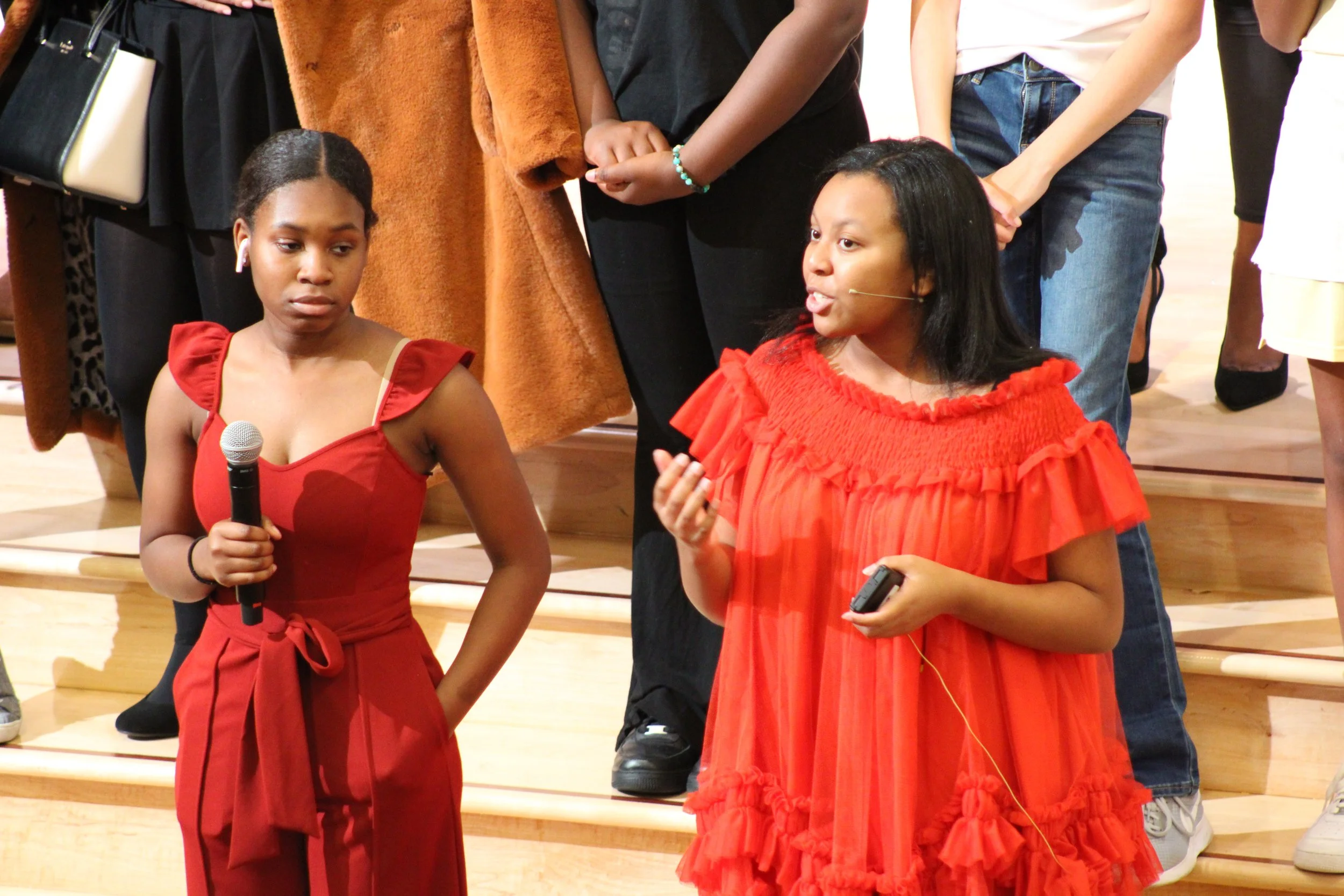 Two women engaging in a conversation on stage, one holding a microphone, with a group of people standing behind them.