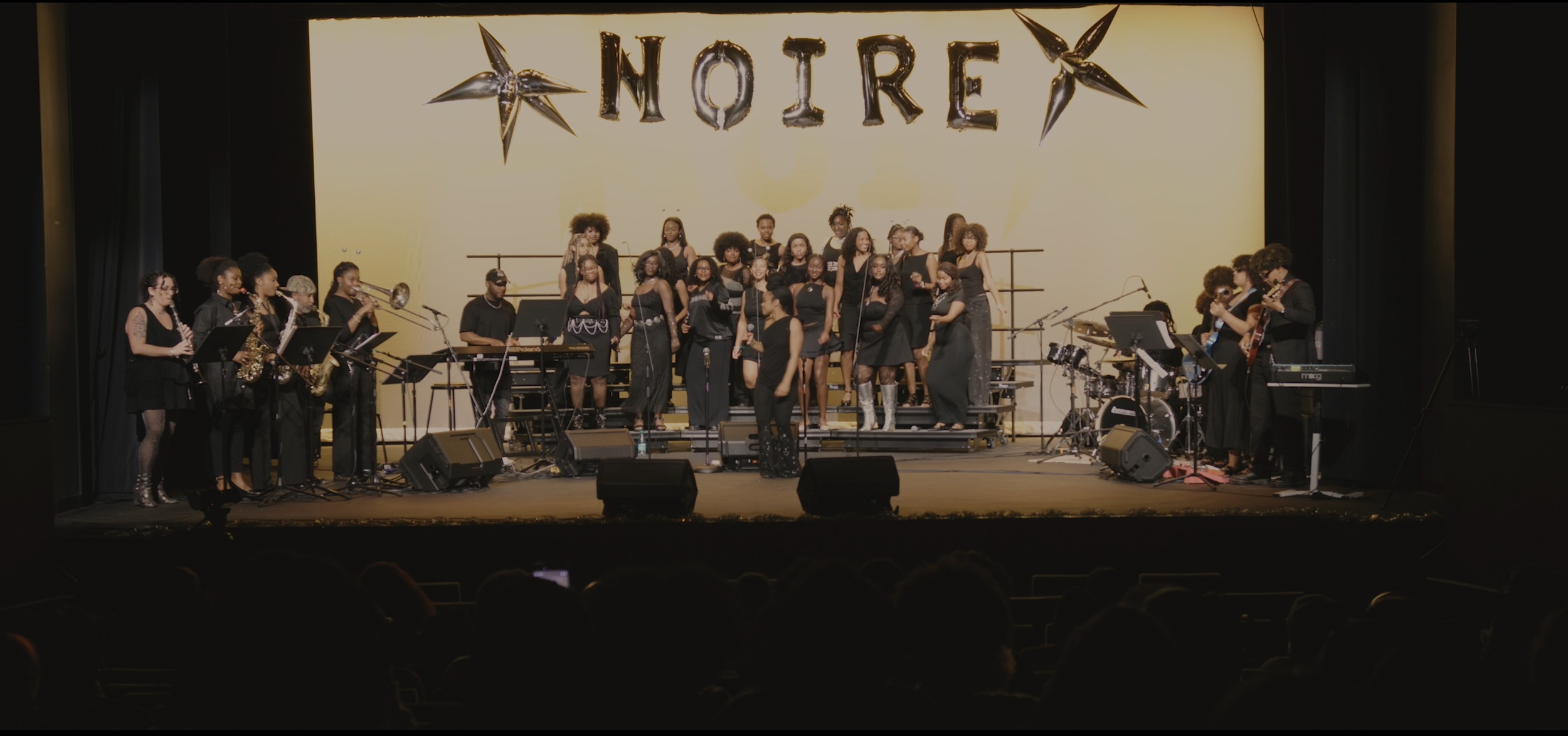 A choir and band performing on stage in front of a large yellow backdrop with the word 'NOIR' spelled out in shiny metallic letters and star-shaped decorations.