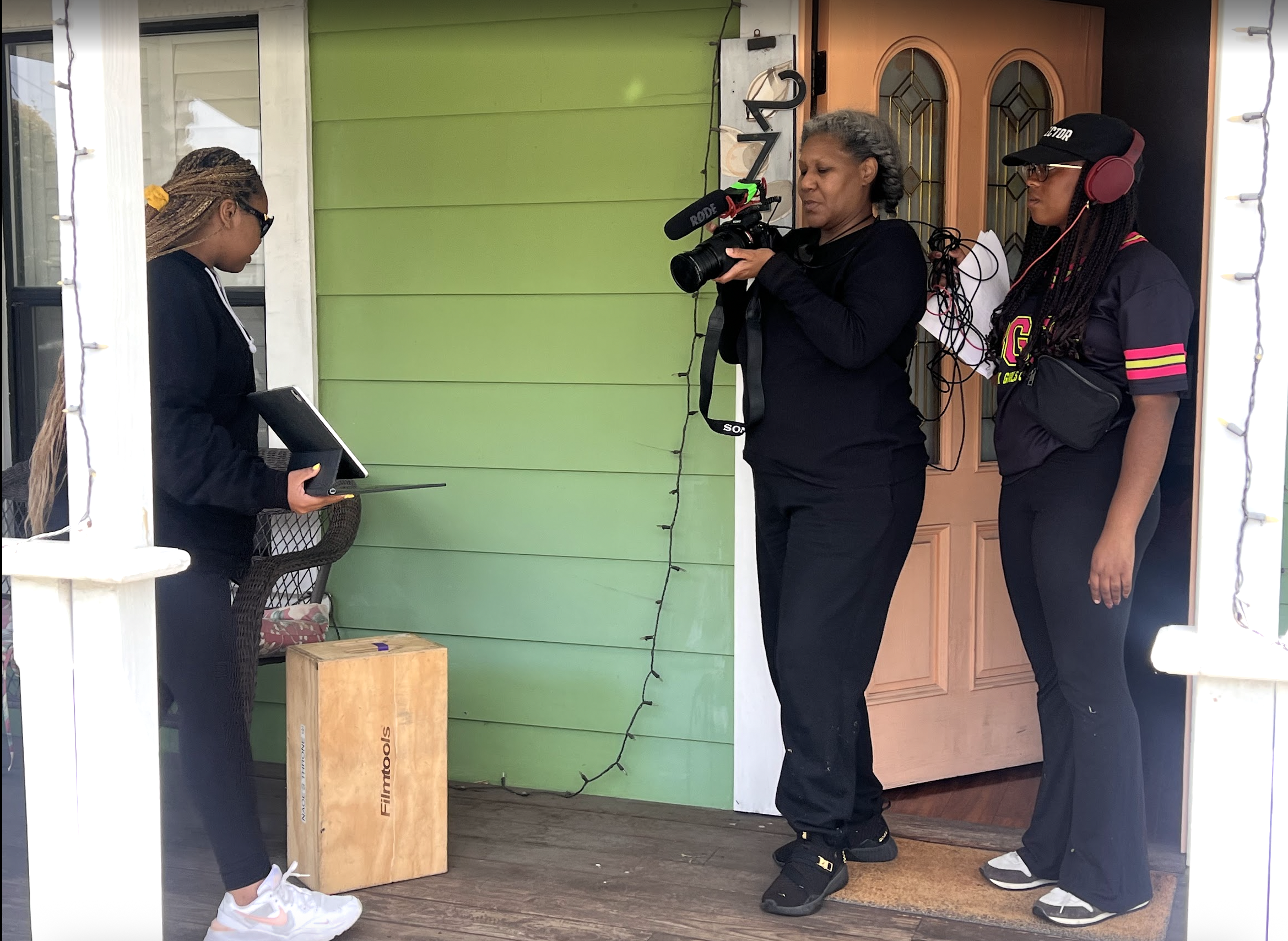 Three women, one holding a camera and two standing nearby with a laptop, are filming a young woman who is standing on a porch holding a notebook. The porch has a green wall and a wooden door.