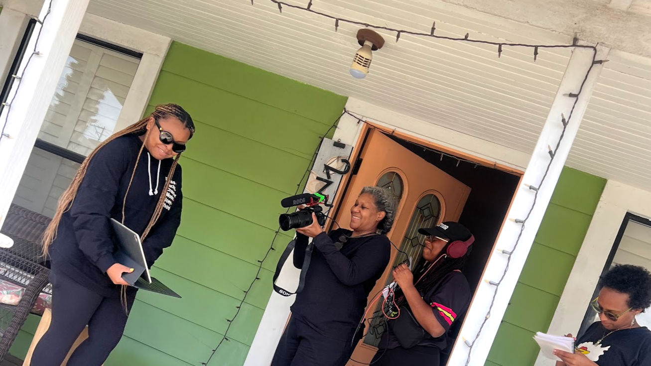 Three women interviewing and recording at a front porch with a lockdown green wall, a door, and string lights.