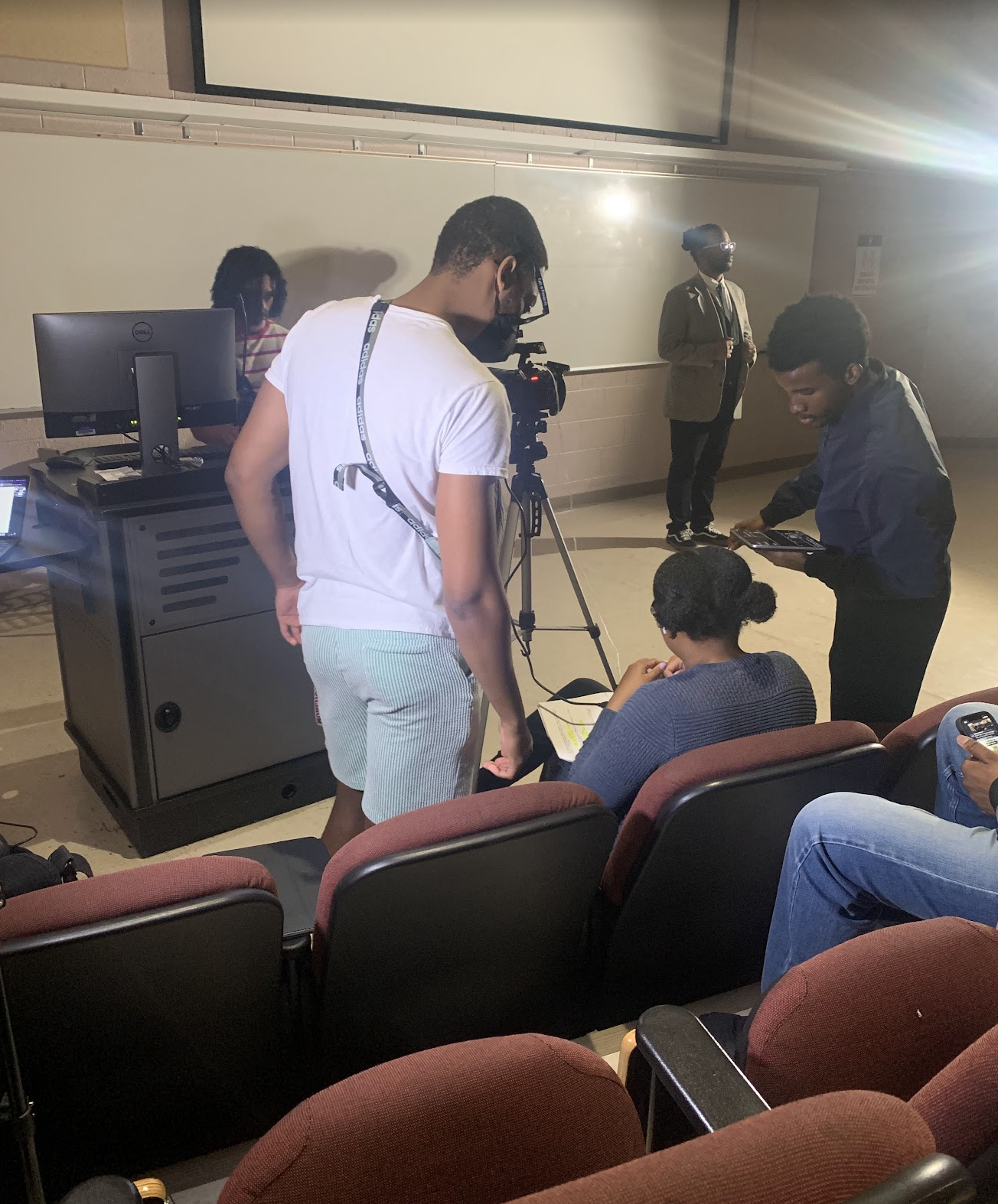 A group of people in a classroom setting, with some preparing technical equipment and others seated in chairs, engaged in what appears to be a filming or presentation activity.