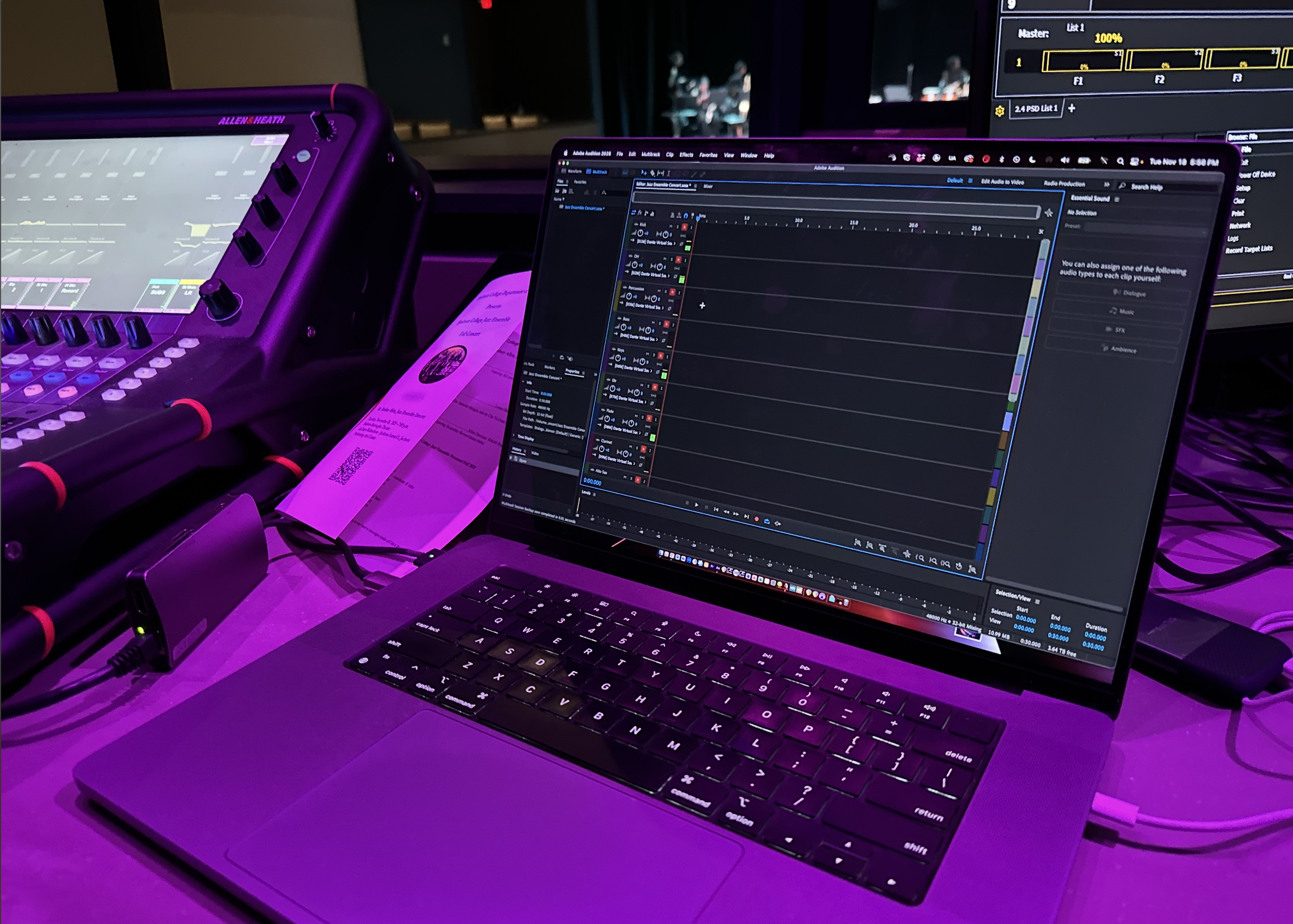 Photo shows a music production setup with a laptop running audio editing software, a control surface with knobs and buttons, a printed receipt, and an audio interface connected via cables. In the background, there are two monitors displaying sound le
