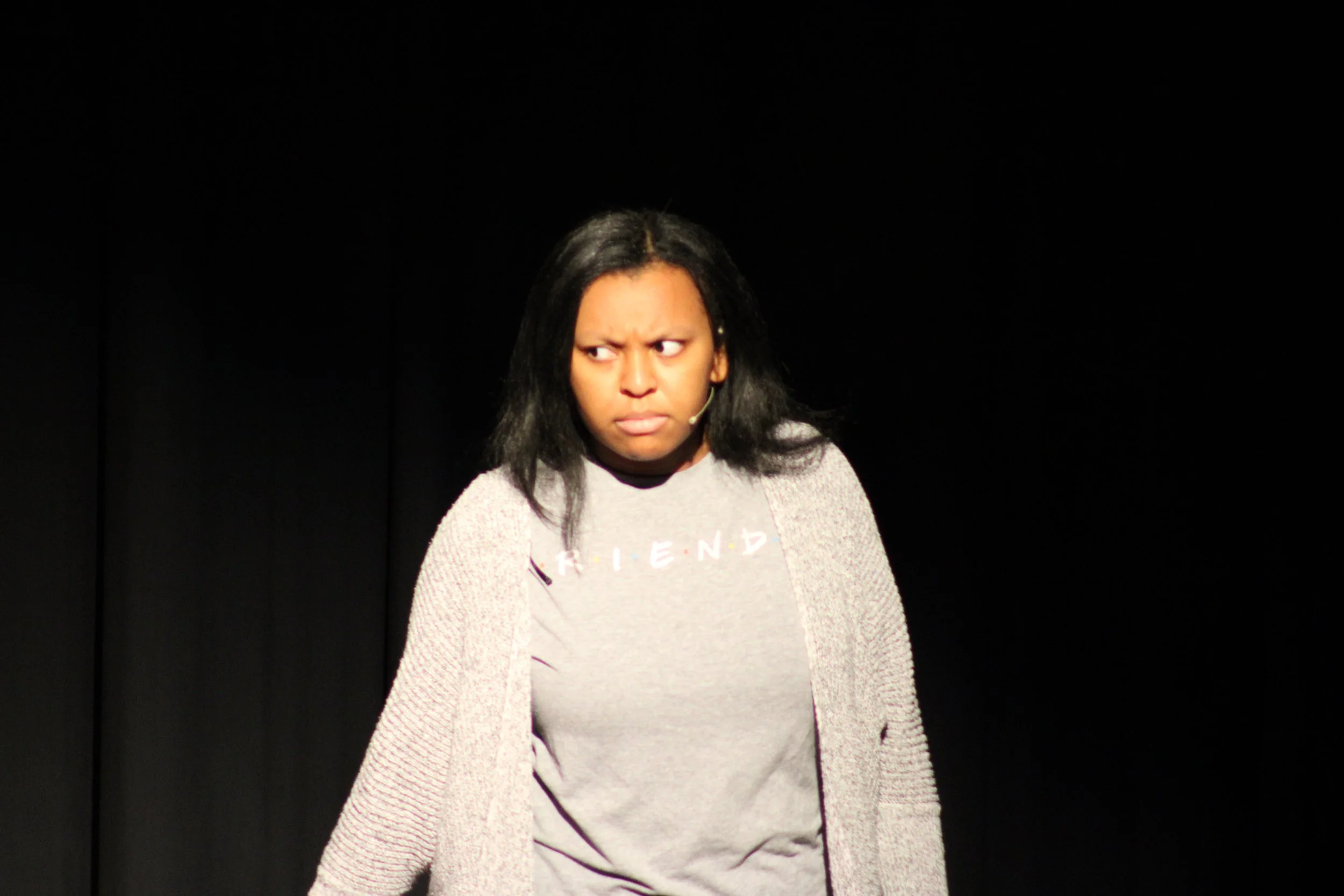 A woman with black hair and skin tone is on stage, looking to her left with a confused or annoyed expression, wearing a gray T-shirt and a light-colored cardigan, with a black background.