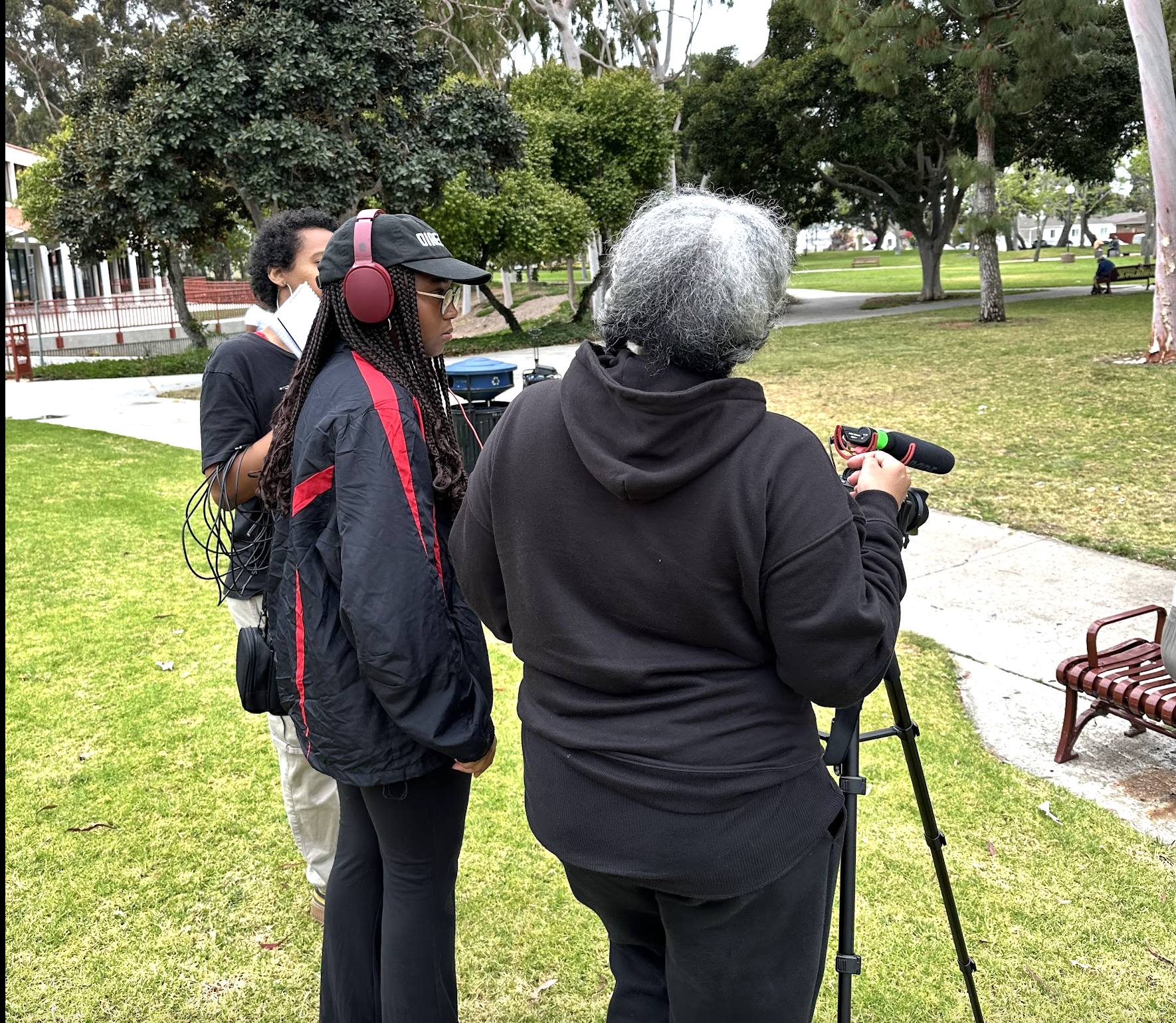 Three people stand outdoors in a park, with two women facing away from the camera and a man with gray hair operating a camera on a tripod, as trees and a sidewalk are visible in the background.