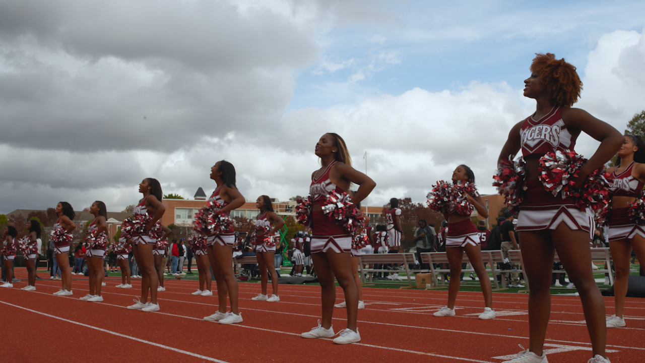 Cheerleaders standing on a track during a sports event, holding pom-poms and wearing matching maroon and white uniforms with 