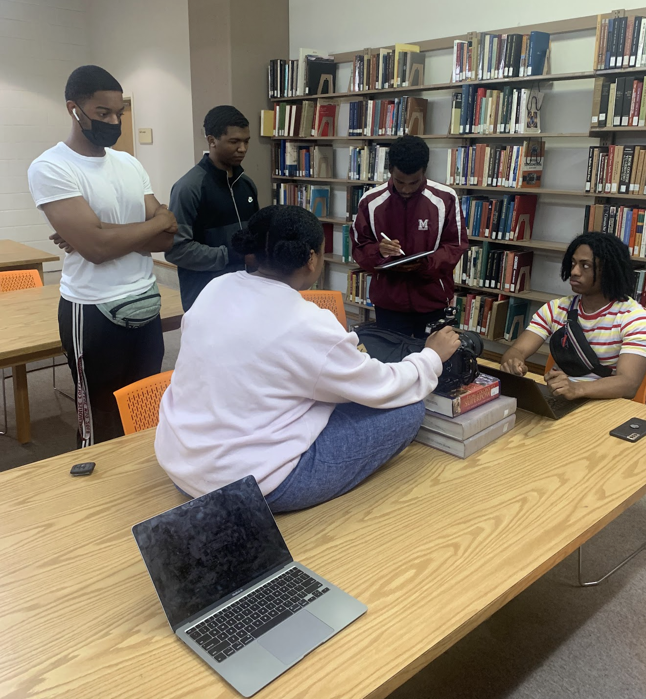 Five young people in a library around a table, with one person recording video, one with a camera, and another with a laptop and notebook, engaging in a group discussion or project.