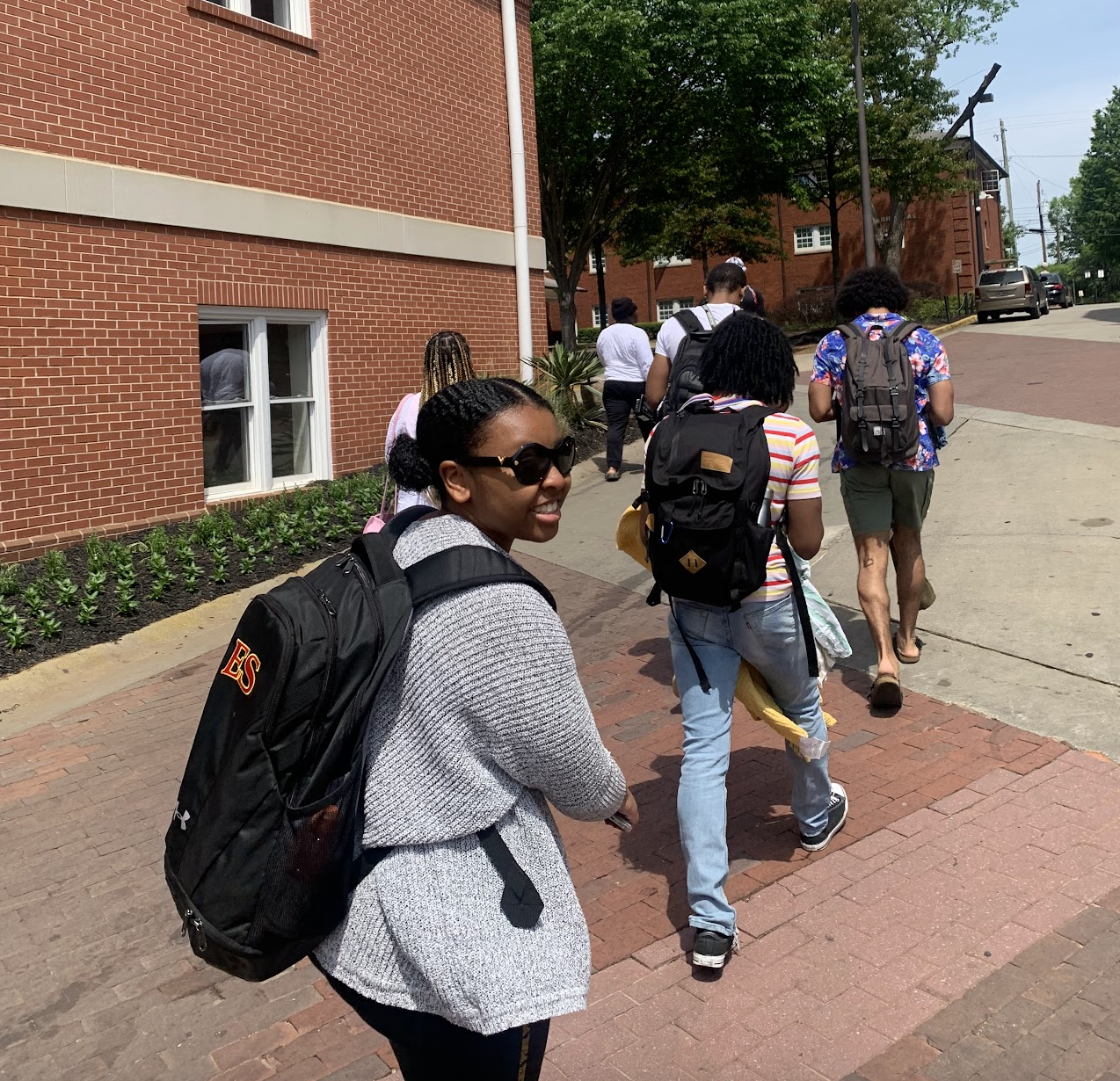 Group of diverse students walking on sidewalk with backpacks, smiling, on a sunny day near a red brick building and trees.
