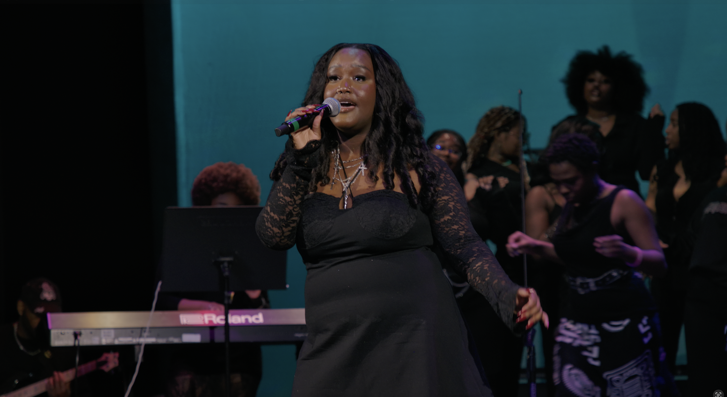 A woman singing into a microphone on stage, surrounded by a group of women and musicians in the background.