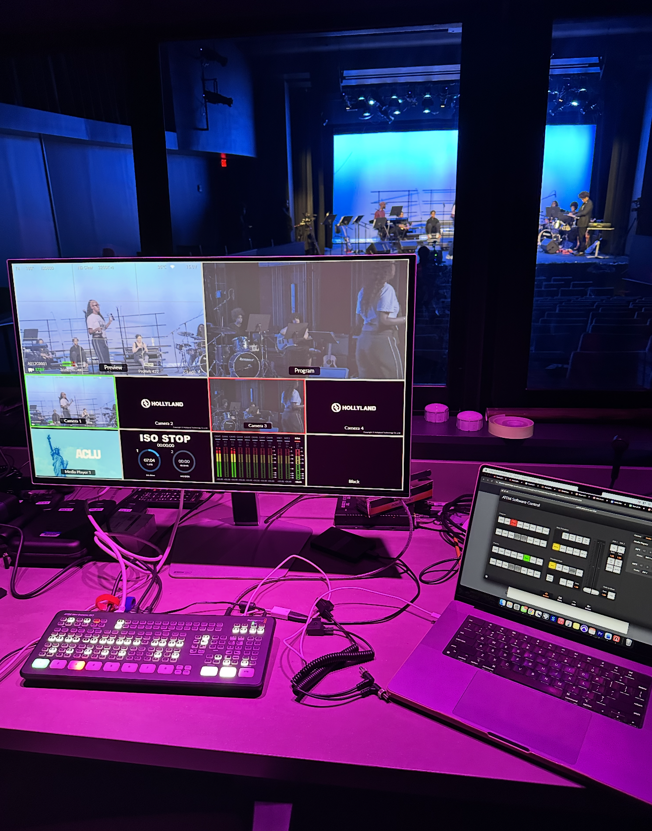 View of a production control station with monitors, a lighting control panel, and a computer, overlooking a stage where a band is rehearsing or performing in a dark auditorium with blue lighting.