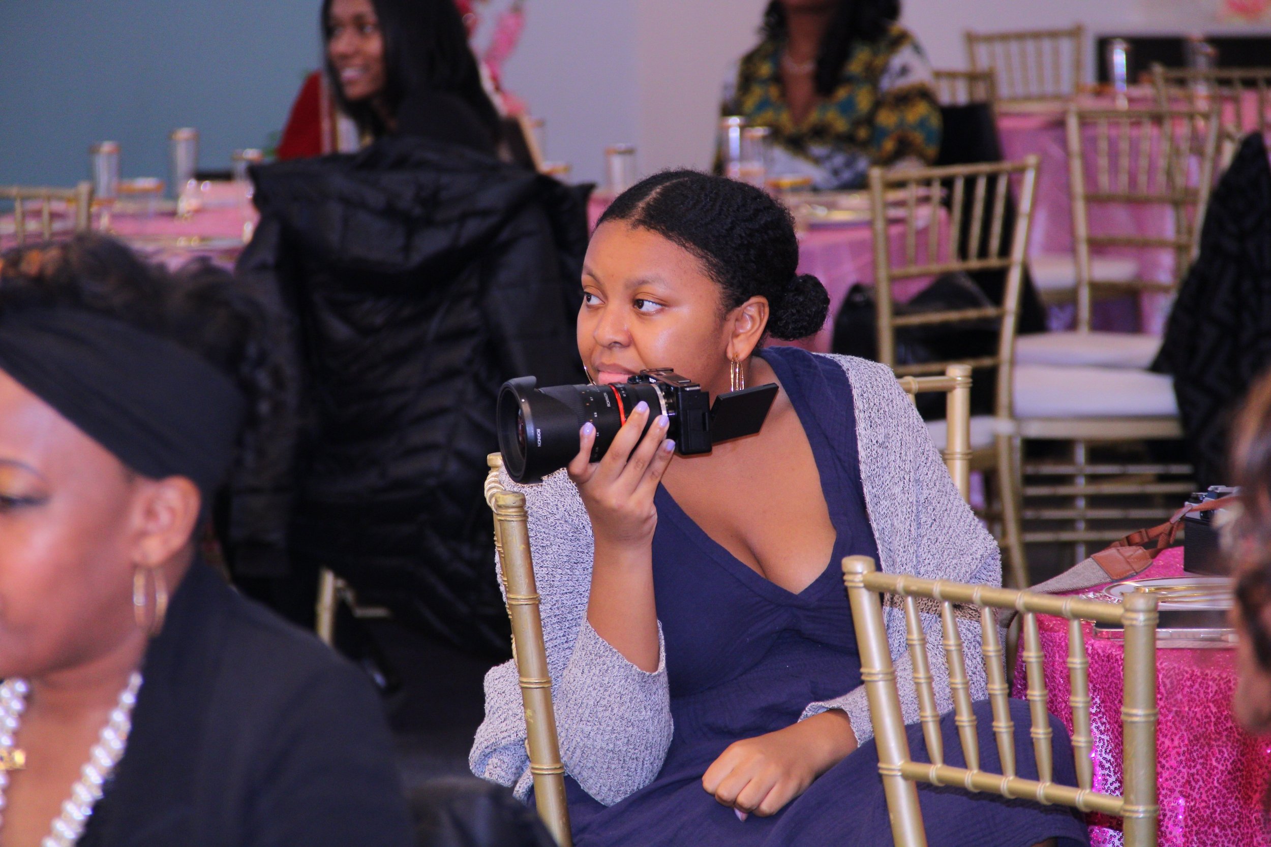 A woman sitting at a table holding a professional camera with a battery grip, looking to her left, at an indoor event with pink tablecloths and gold chairs, other women are visible in the background.