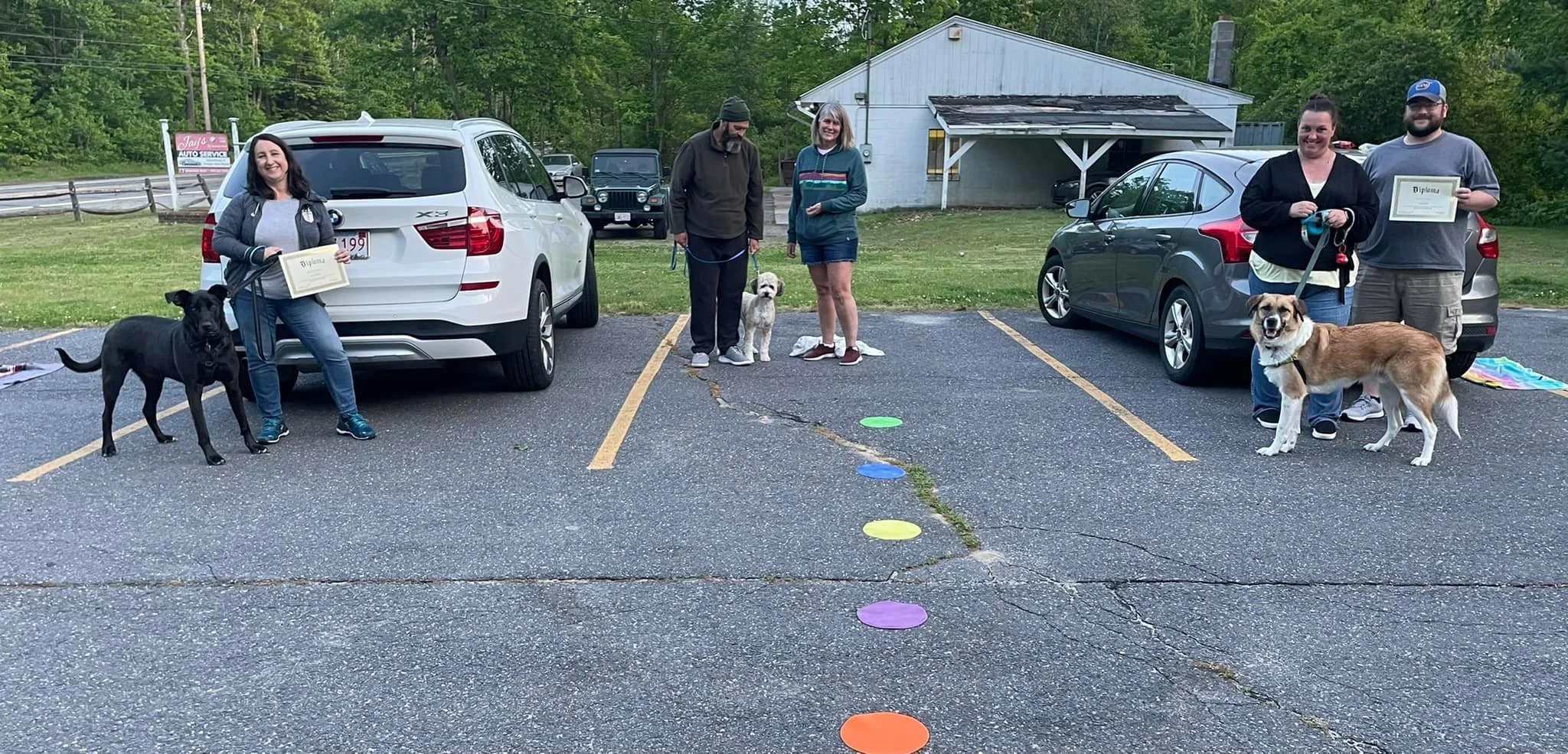 People gathered in a parking lot with their dogs, holding certificates, near two parked cars with a small building and trees in the background.