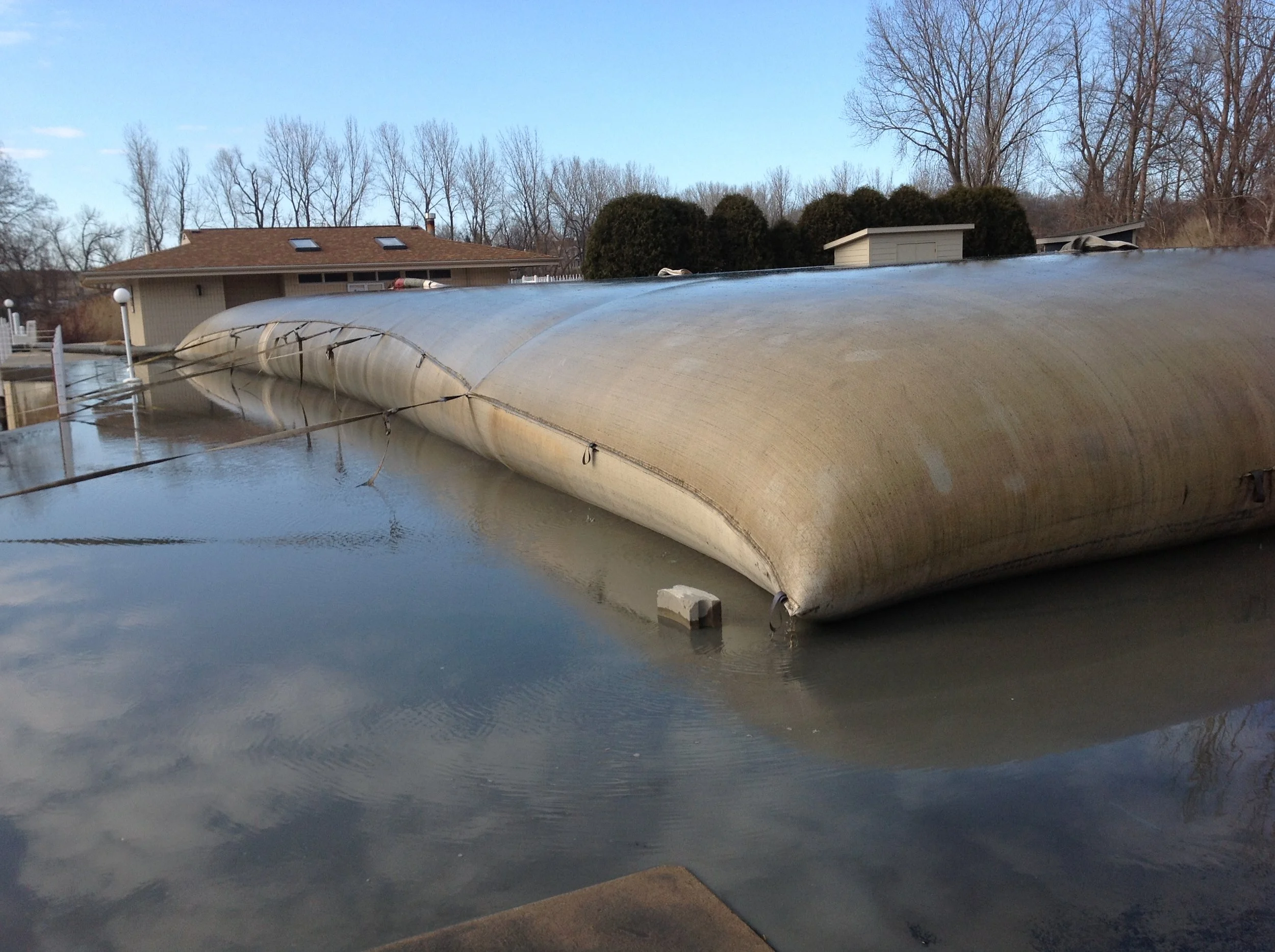A large, closed boat covered with a tarp resting on the water's surface at a dock, with houses and leafless trees in the background on a clear day.