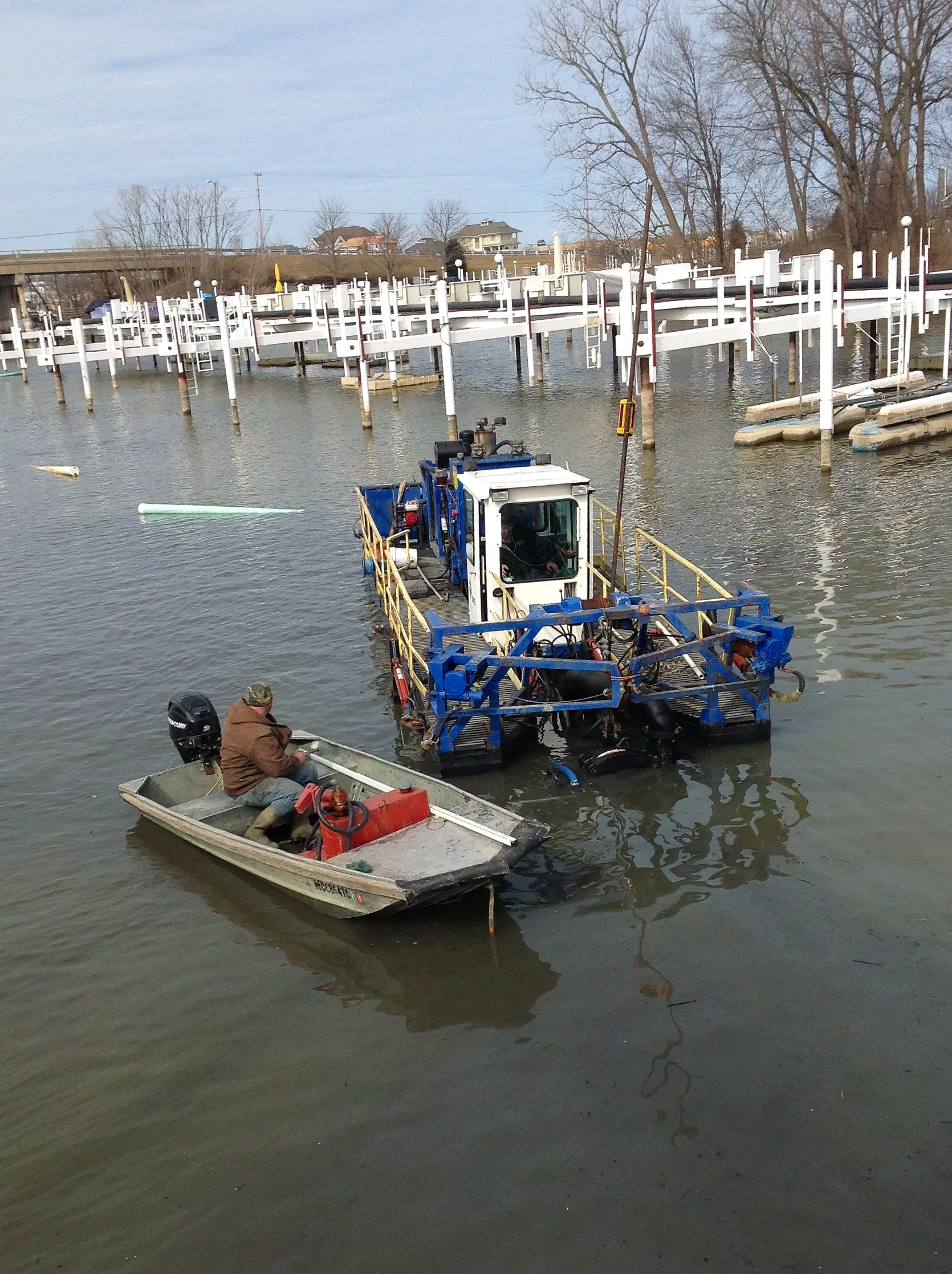 A man in a small boat operating a blue underwater inspection vehicle on a river with boat docks and empty floating boat slips in the background.