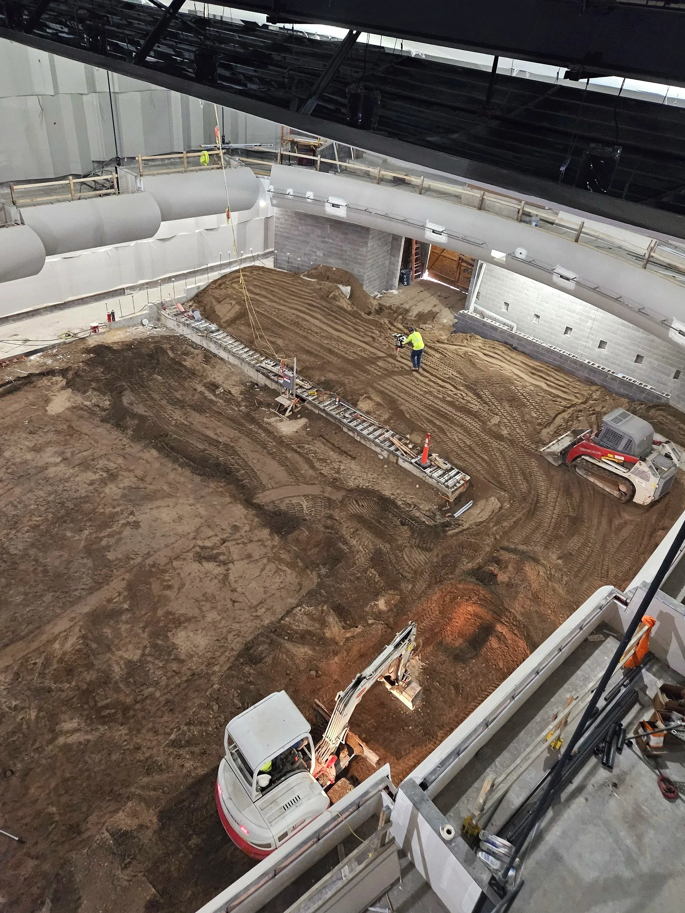 Construction site inside a building with worker and machinery, dirt and gravel ground, temporary walls, and large ventilation pipes.