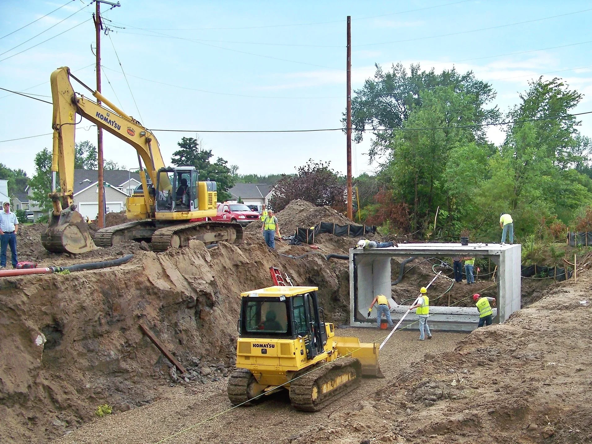 Construction workers and heavy machinery building a bridge or tunnel entrance in a dirt excavation site, with trees and residential houses in the background.