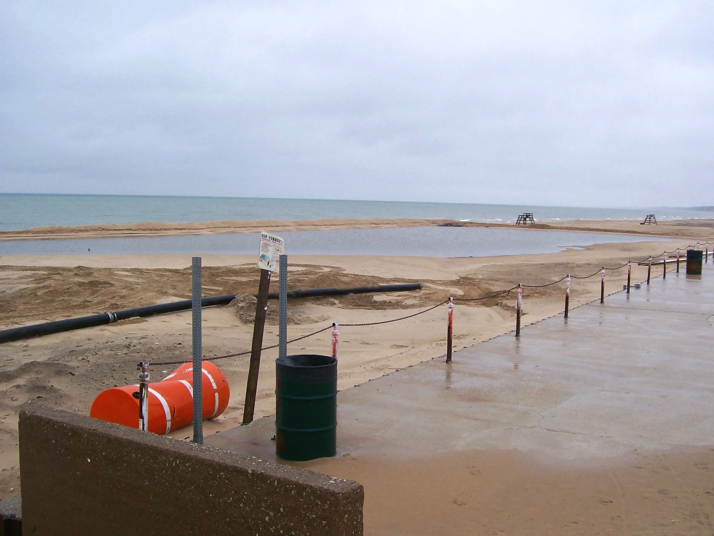 A beach scene on a cloudy day with a sandy shoreline, a small body of water parallel to the ocean in the background, and lifeguard chairs on the distant sand dunes. A black pipe and an orange life-preserver are on the concrete walkway, which is separ