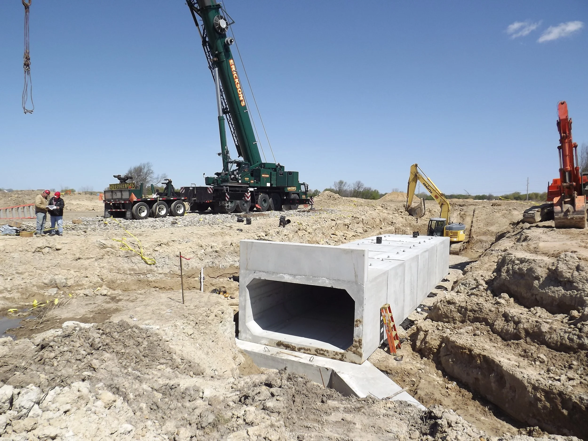 Construction site with large green crane, yellow excavator, and orange machinery, showing workers and concrete drainage pipe installation.
