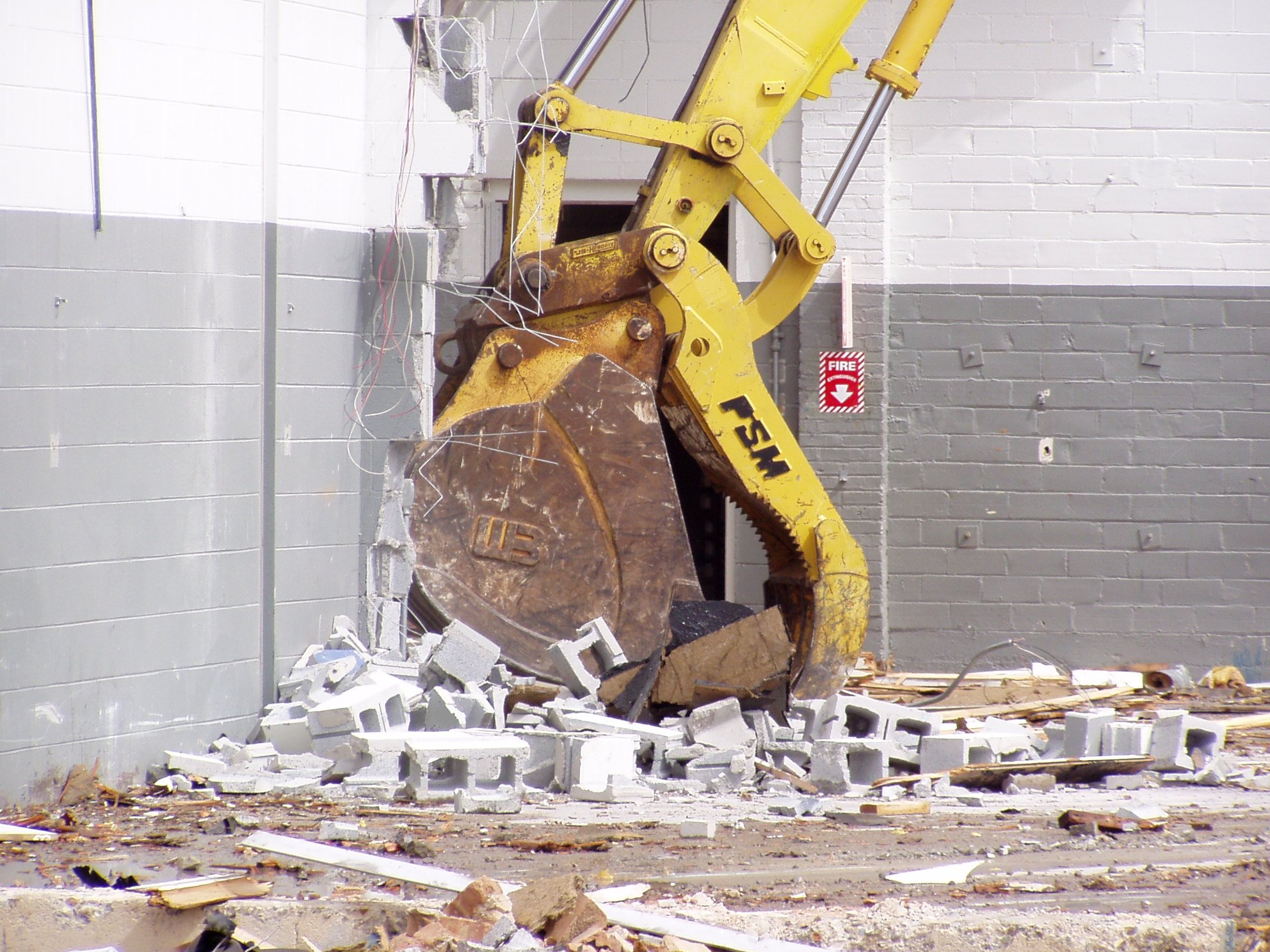 A yellow hydraulic breaker demolishing a wall made of concrete blocks inside a room, with debris scattered on the floor.