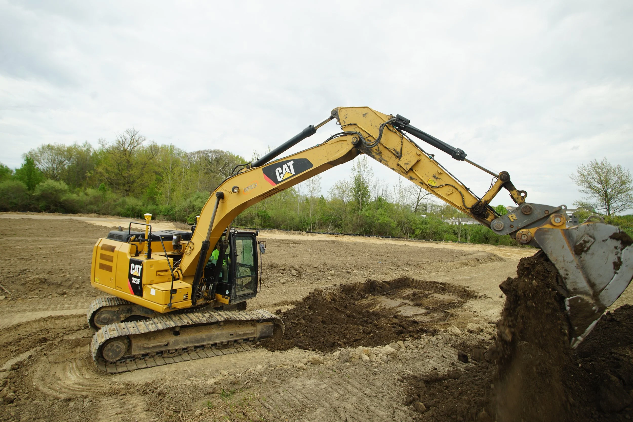 A yellow Caterpillar excavator digging into the ground on a construction site with trees and overcast sky in the background.