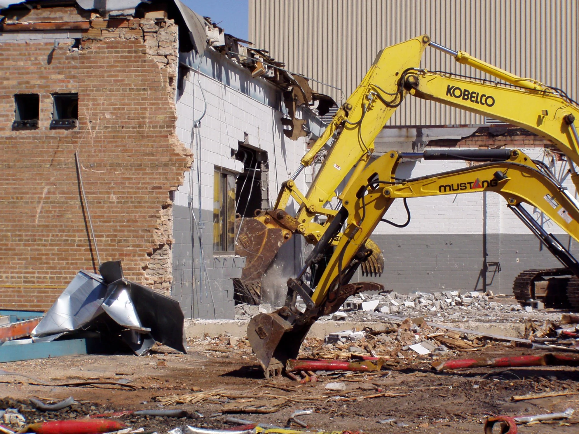 Two yellow construction excavators demolishing a brick and concrete building, with debris and rubble on the ground.