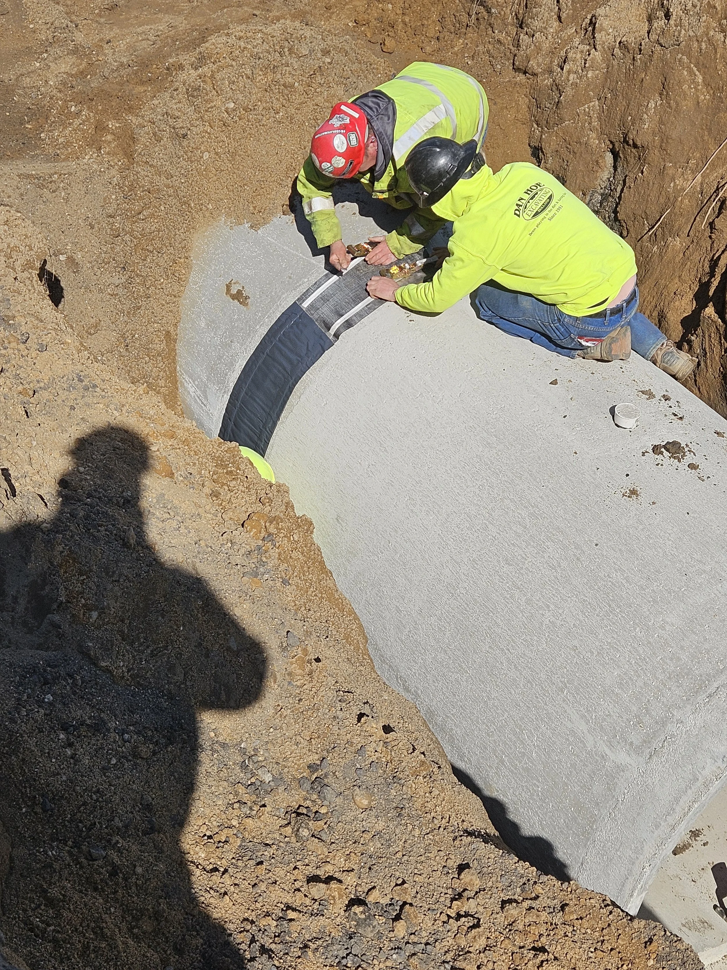 Two construction workers in safety gear installing a large concrete pipe underground.