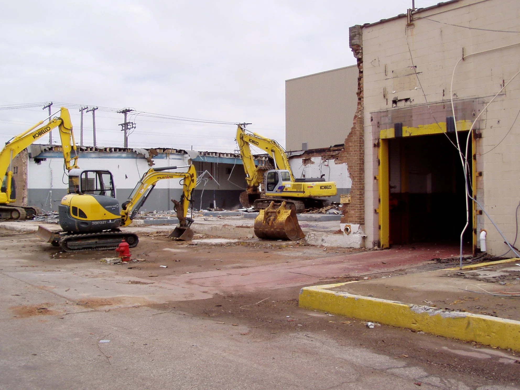 Construction site with three yellow excavators demolishing a building, debris on the ground, and a partially dismantled structure with exposed bricks and wires.