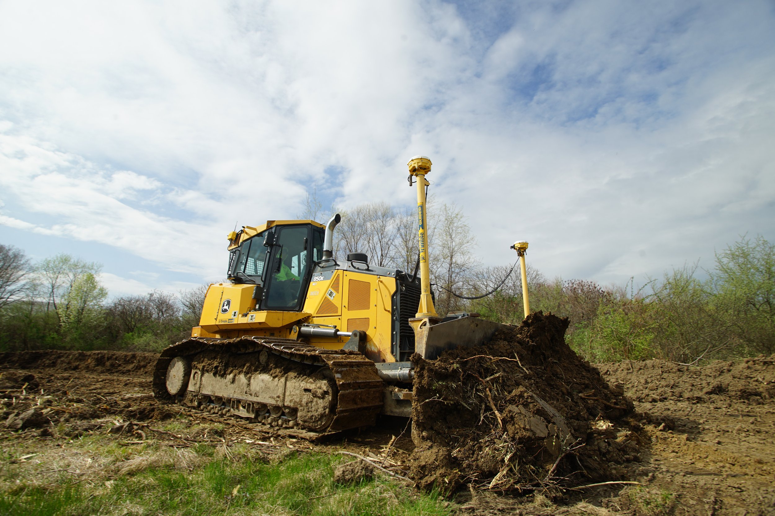 Yellow bulldozer working on a construction site with a large pile of dirt, trees, and a cloudy sky in the background.