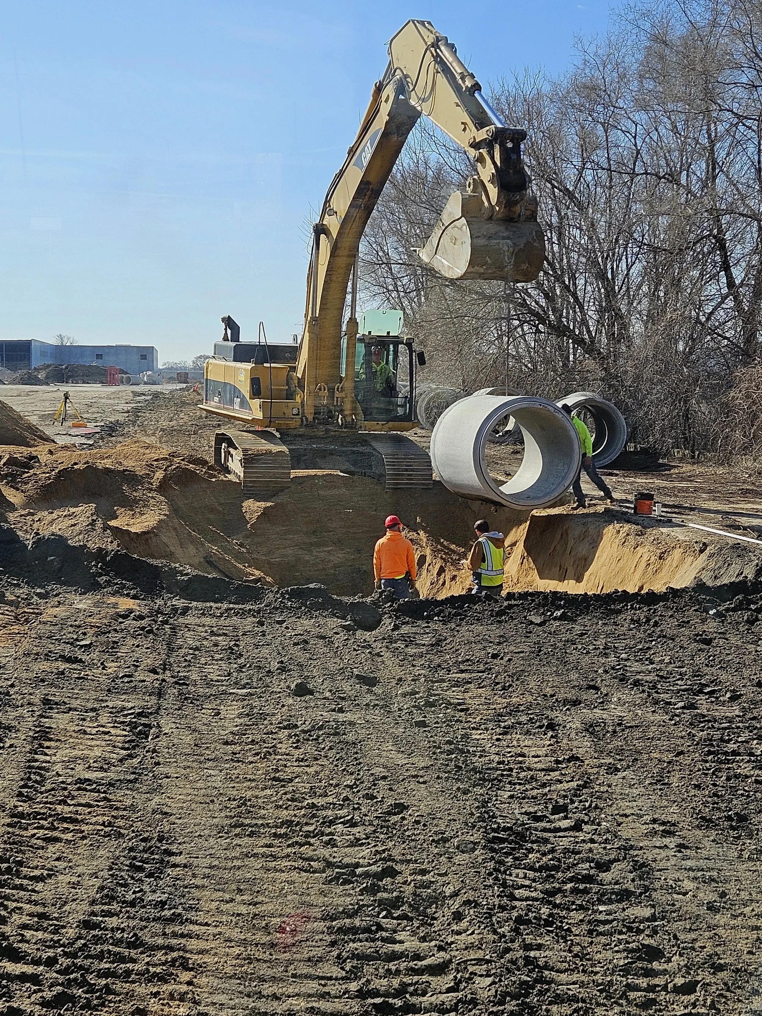 Construction site with a large excavator lifting a concrete pipe, workers in safety vests and helmets work nearby, and there are trees in the background.
