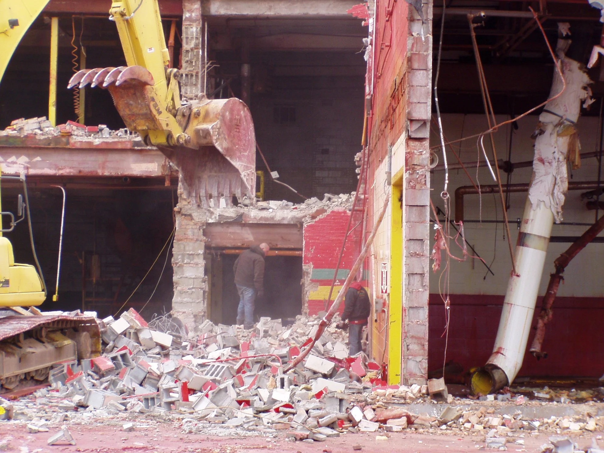 Construction site with a yellow excavator demolishing a brick building, debris scattered on the ground, and two workers in the background.